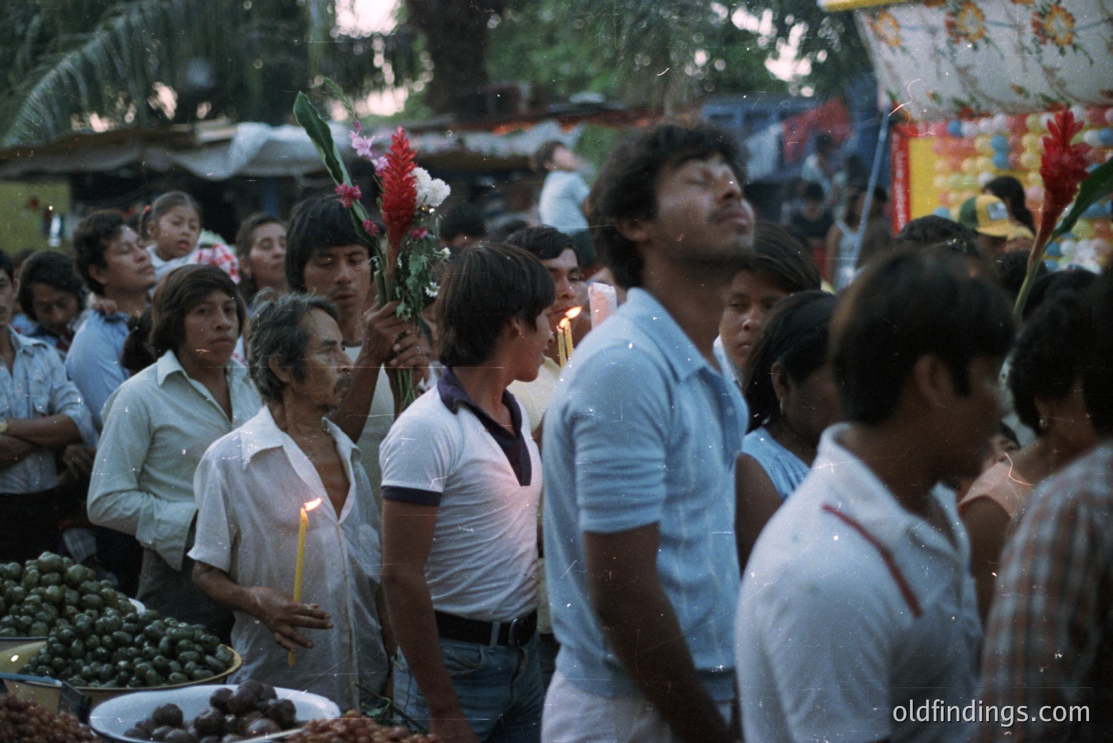 A candid street scene shows a crowd of people during what appears to be a religious procession. Numerous individuals hold lit candles, while others carry vibrant floral arrangements. The attire and hairstyles suggest a 1970s timeframe, possibly in Southeast Asia. A market display with fruits is visible.
