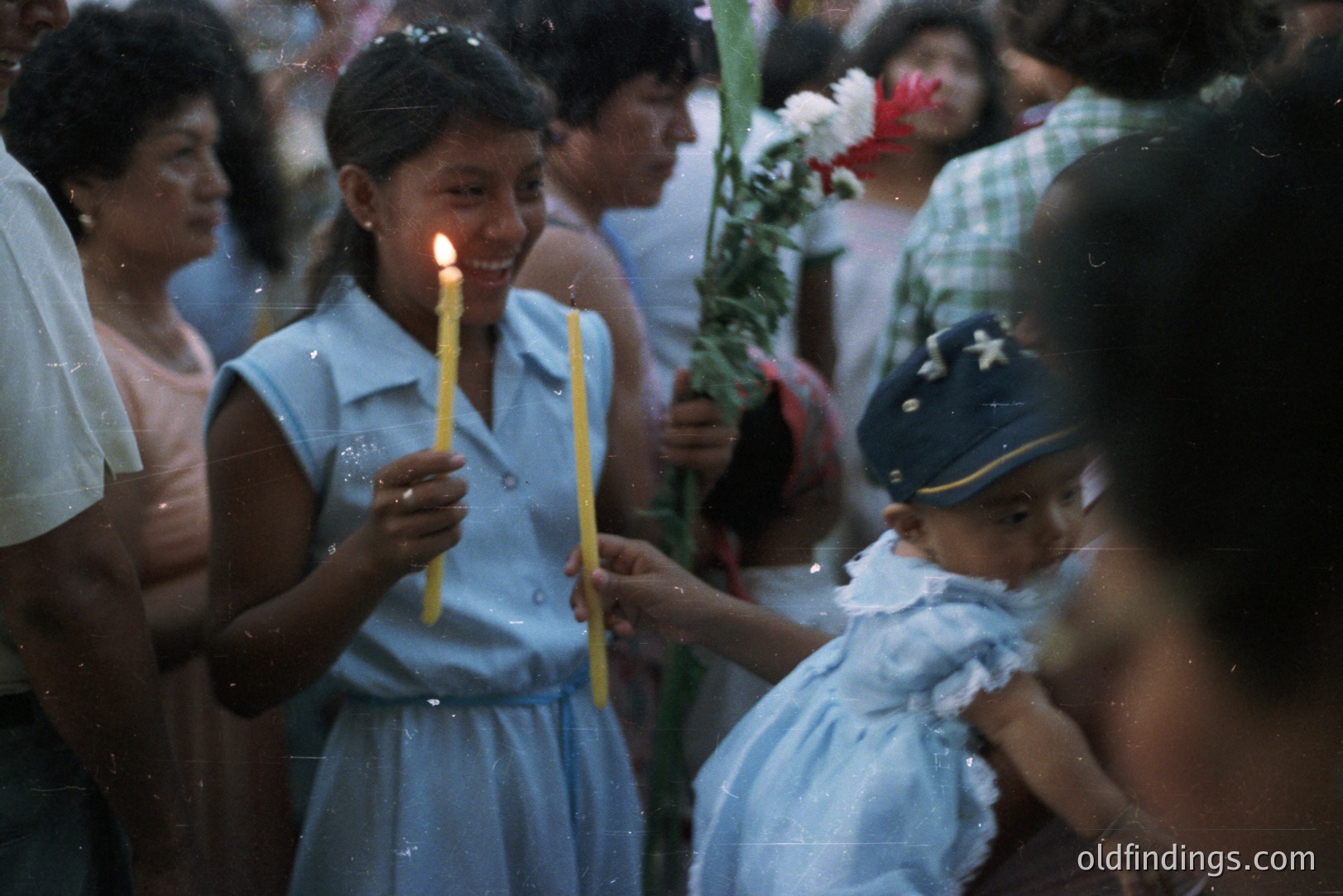 A young girl in a light blue dress and cap holds a bouquet of flowers while a teenage girl, also in blue, carries two lit candles. The image appears to be from a cultural or religious gathering, possibly a procession. Likely taken in the 1970s or 80s, judging by the clothing and film quality. The scene conveys a sense of community and tradition.