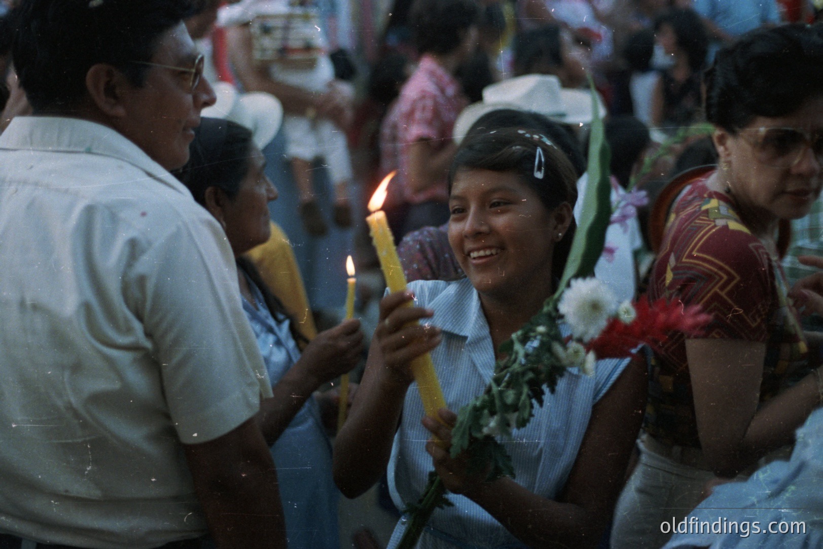 A group gathers in what appears to be a festive, outdoor scene. A young woman holds a lit candle, smiling warmly, surrounded by people; a man and another woman are visible nearby. The image features a vintage aesthetic, likely dating to the 1970s. The clothing styles suggest a Latin American setting. The blurred background indicates movement and a crowded atmosphere.