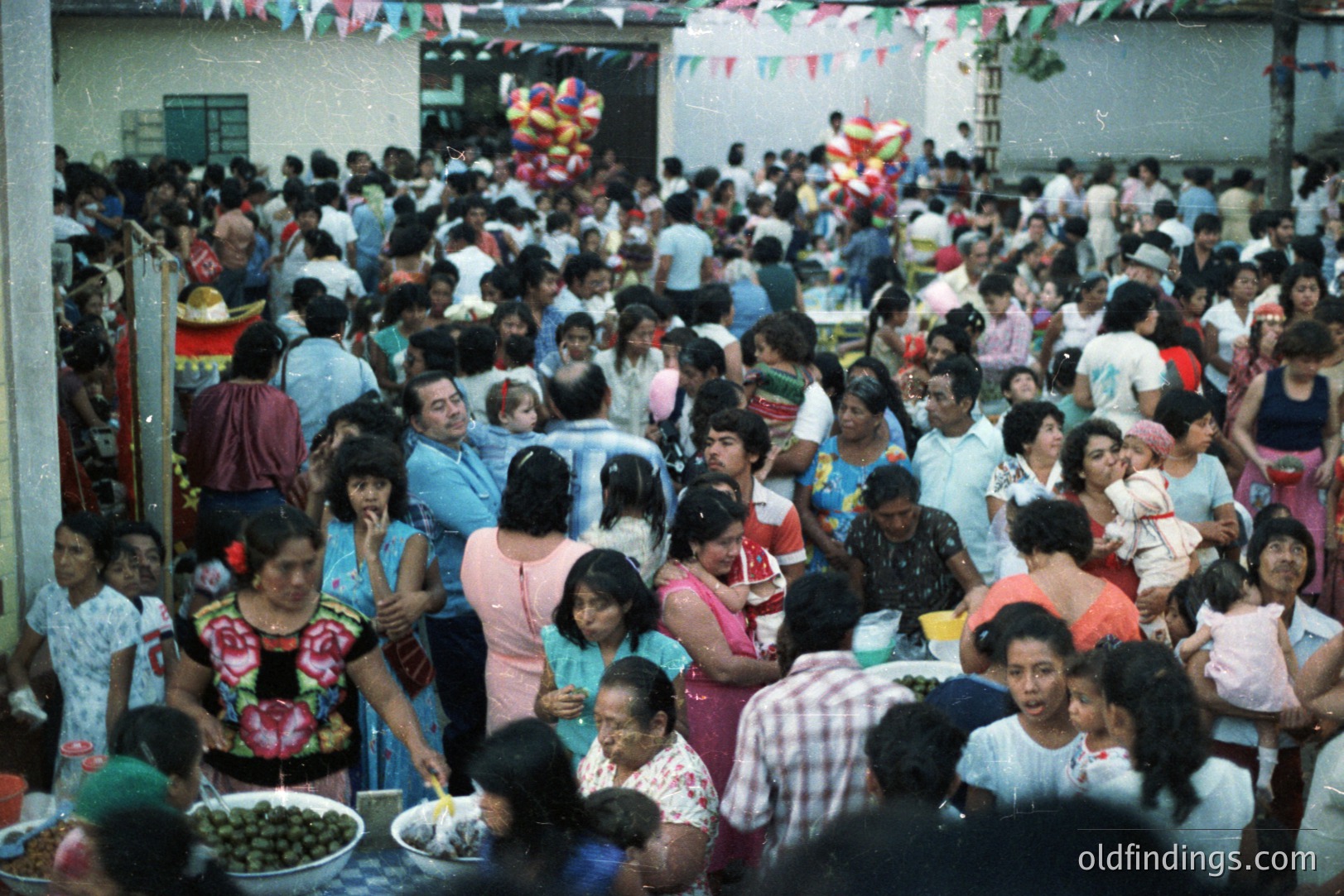 Crowded outdoor gathering, possibly a festival or community event. Numerous people of various ages are present, enjoying food and socializing. Decorations of colorful paper chains adorn the background. Likely 1970s-1980s, based on clothing styles. A candid moment of vibrant cultural exchange.