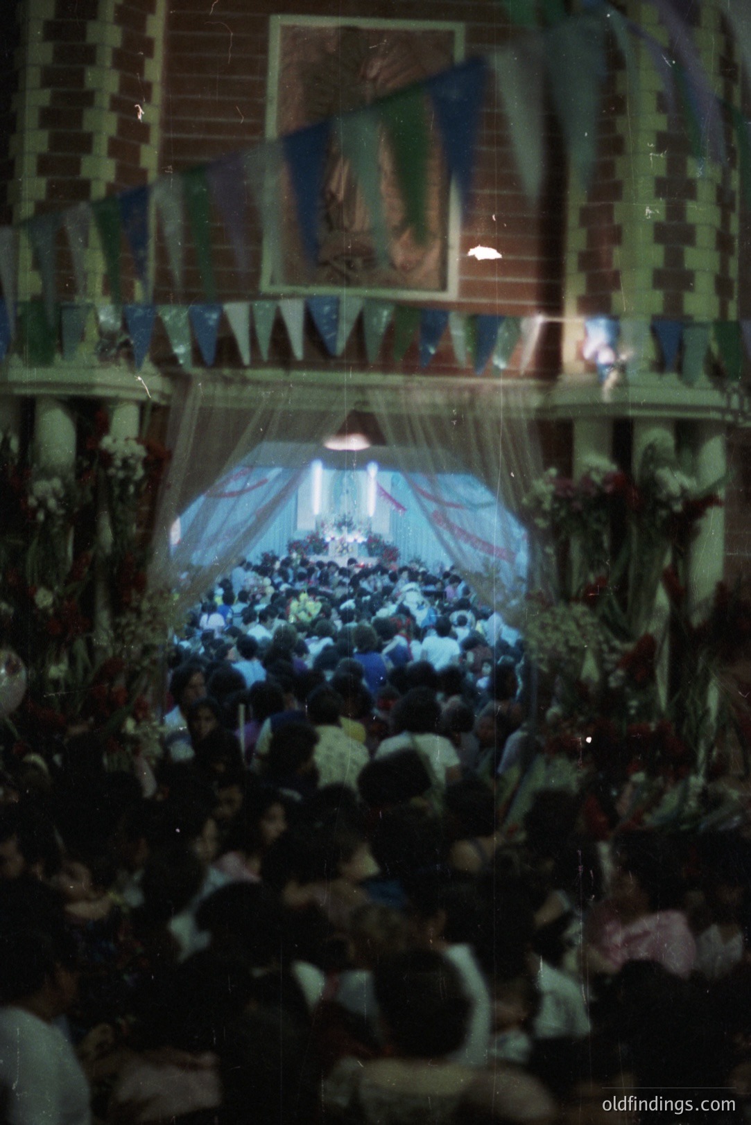 Large crowd gathers beneath decorative flags and floral arrangements, likely for a religious festival or community celebration. Image exhibits a slightly grainy aesthetic typical of 1970s or 80s photography. Festive and bustling atmosphere. Location possibly Southeast Asia.