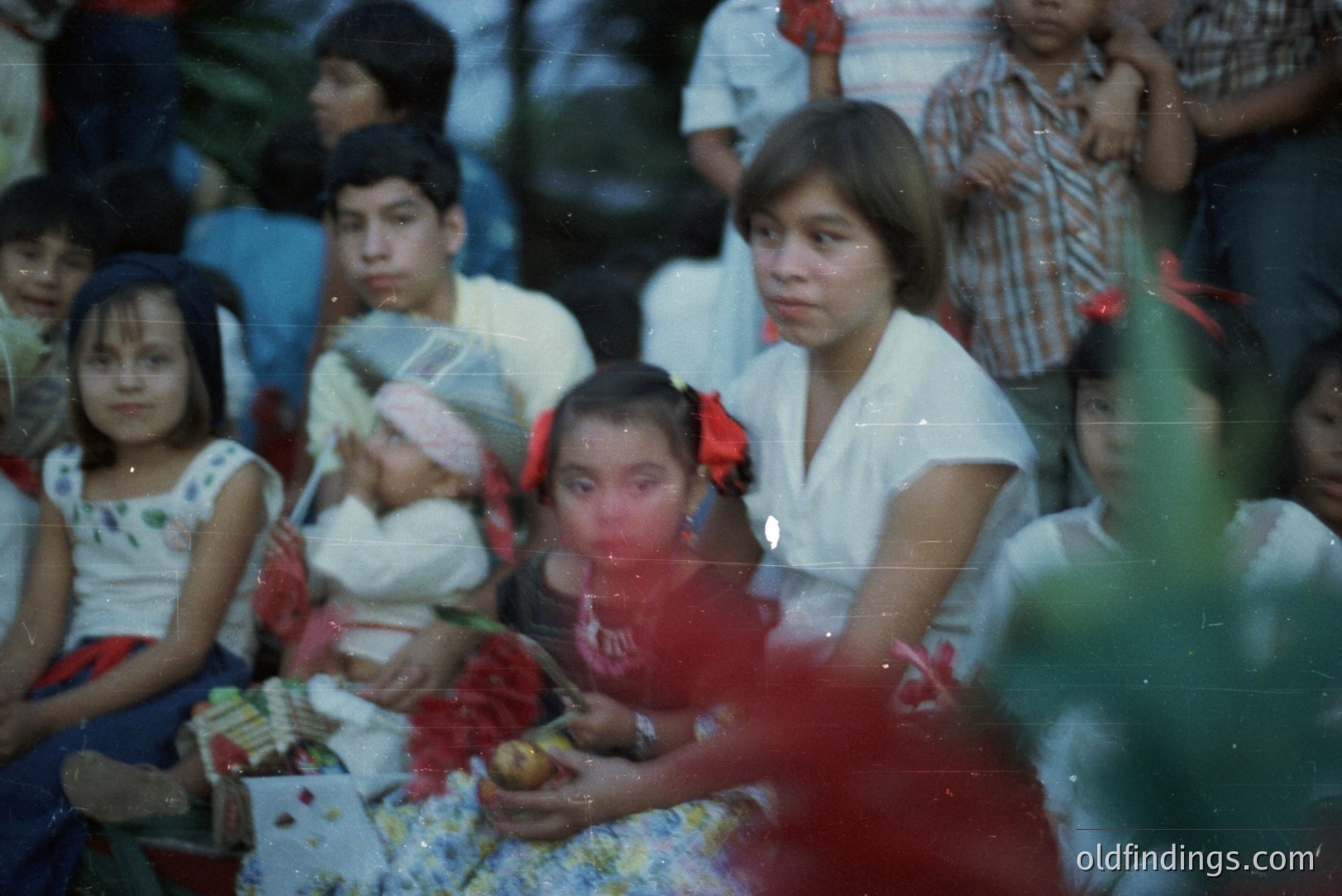 Group portrait shows children, likely in a rural setting. A young girl holds flowers, while a teen holds her hand. Garments suggest a cultural tradition; the photo’s faded colors imply a 1970s timeframe. Subject matter useful for documentary or anthropological research.