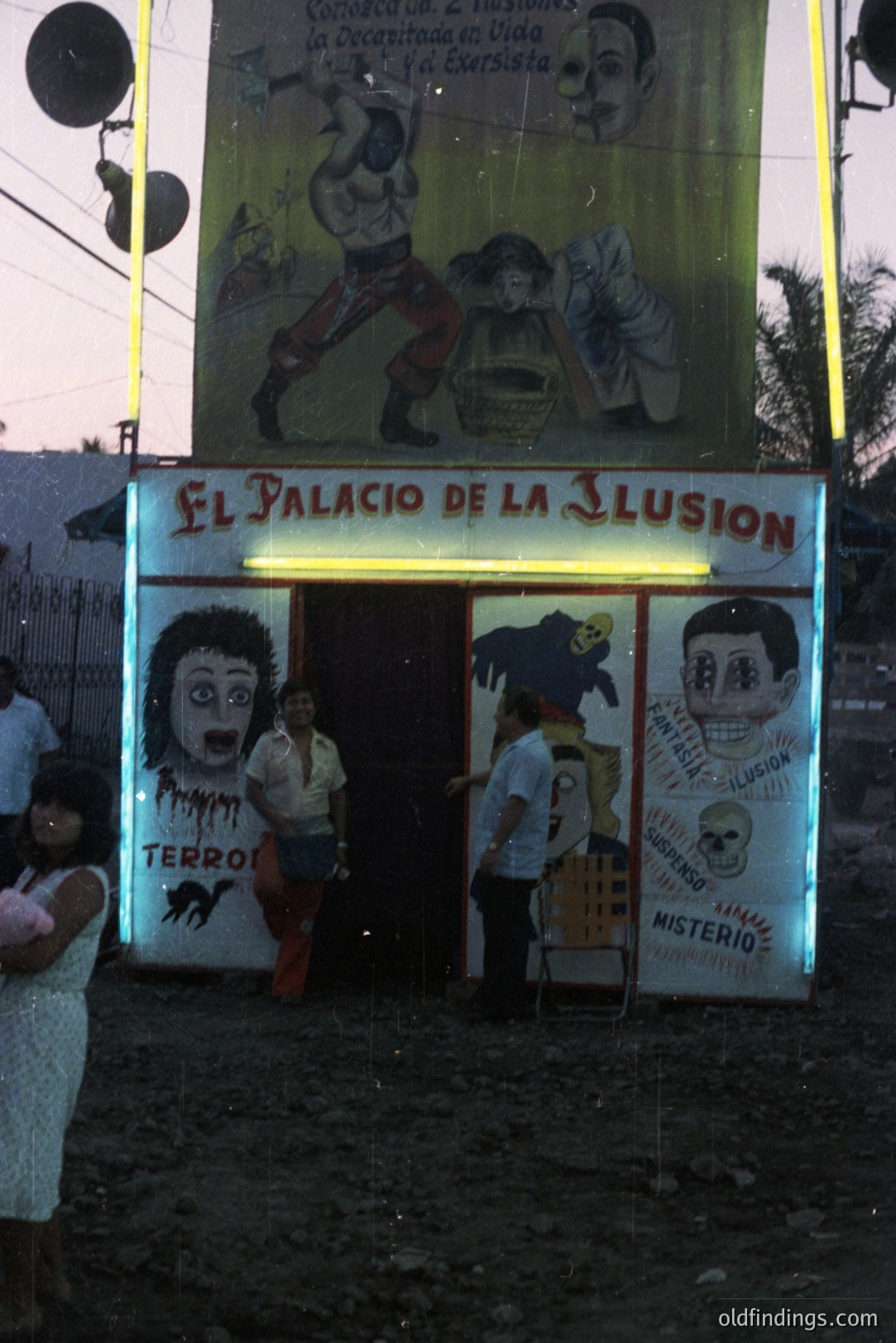 A vibrant, hand-painted sign for "El Palacio de la Ilusion" (Palace of Illusion) advertises a sideshow attraction. The sign features portraits, illustrated illusions, and bold lettering. Two figures are entering the attraction, and a blurred crowd is visible. Likely a tourist destination.
