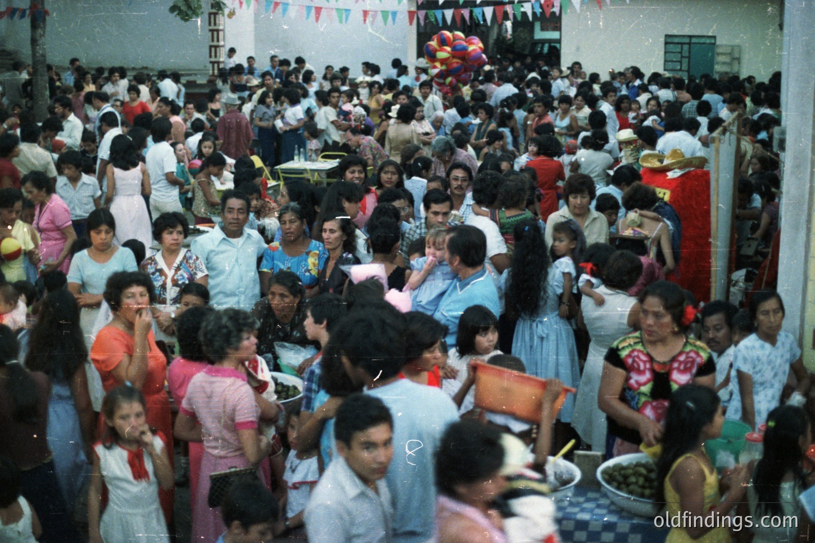 Large crowd gathered outdoors, possibly a festival or community event. People dressed in 1970s fashion—floral prints, pastel dresses, short hairstyles. Banners and balloons adorn the space. Tables display food, likely olives or pickles. Possible Latin American setting.