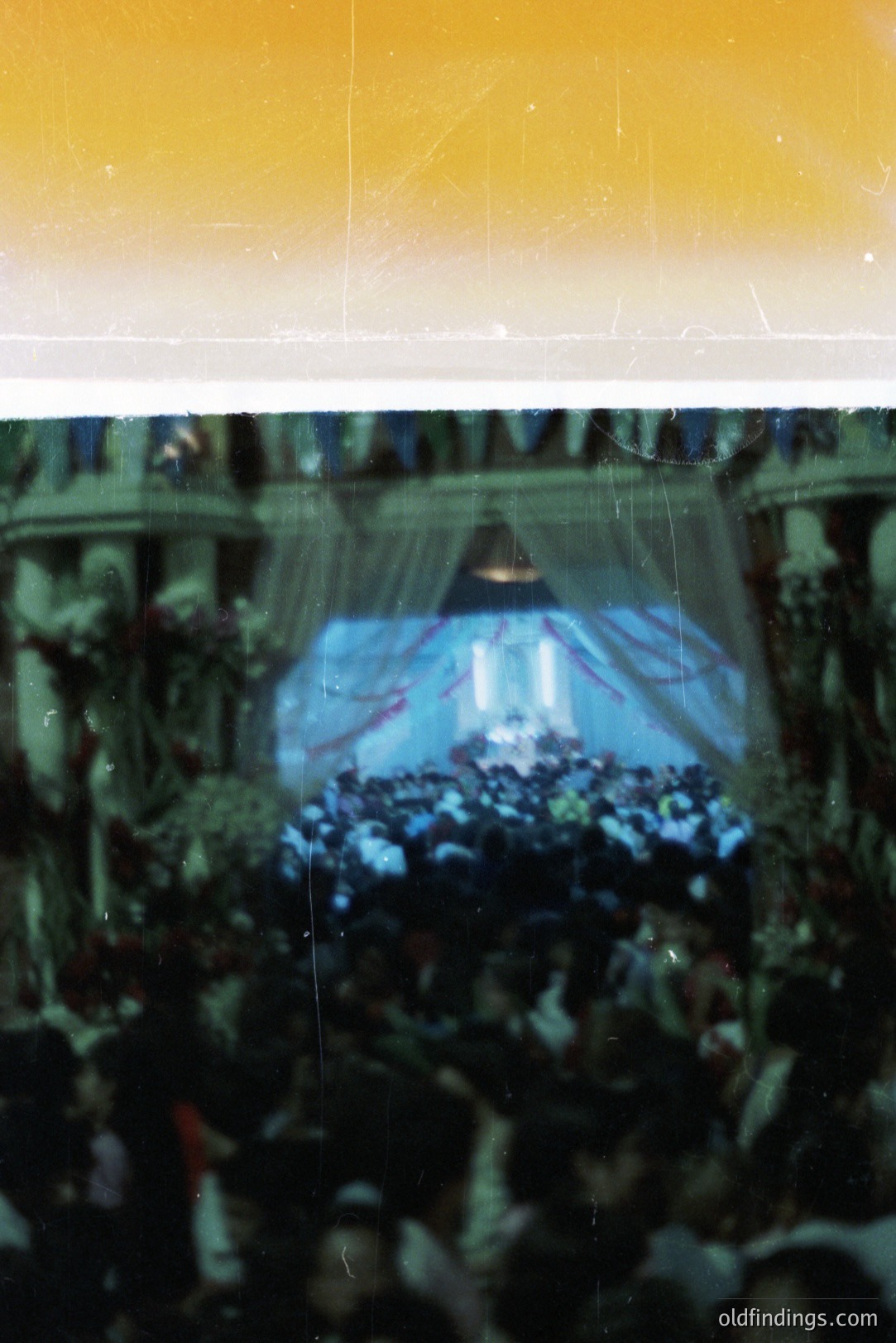 Large crowd gathered before an elaborately decorated stage with draped curtains and floral arrangements. The event appears to be a formal gathering, possibly a concert or public appearance. Warm color cast indicates age or film type.