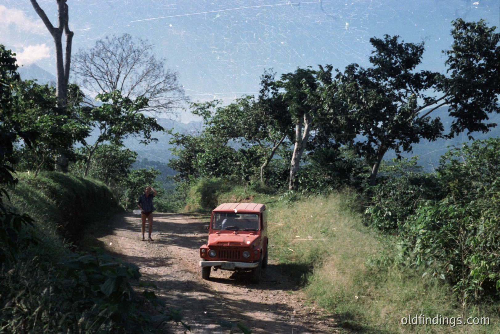 Dusty, unpaved road ascends through dense, tropical foliage. A small, red utility vehicle travels uphill; a lone figure walks away from the viewer. Mountainous landscape with distant peaks visible. Likely taken in the 1970s-80s, possibly Southeast Asia or Central America.