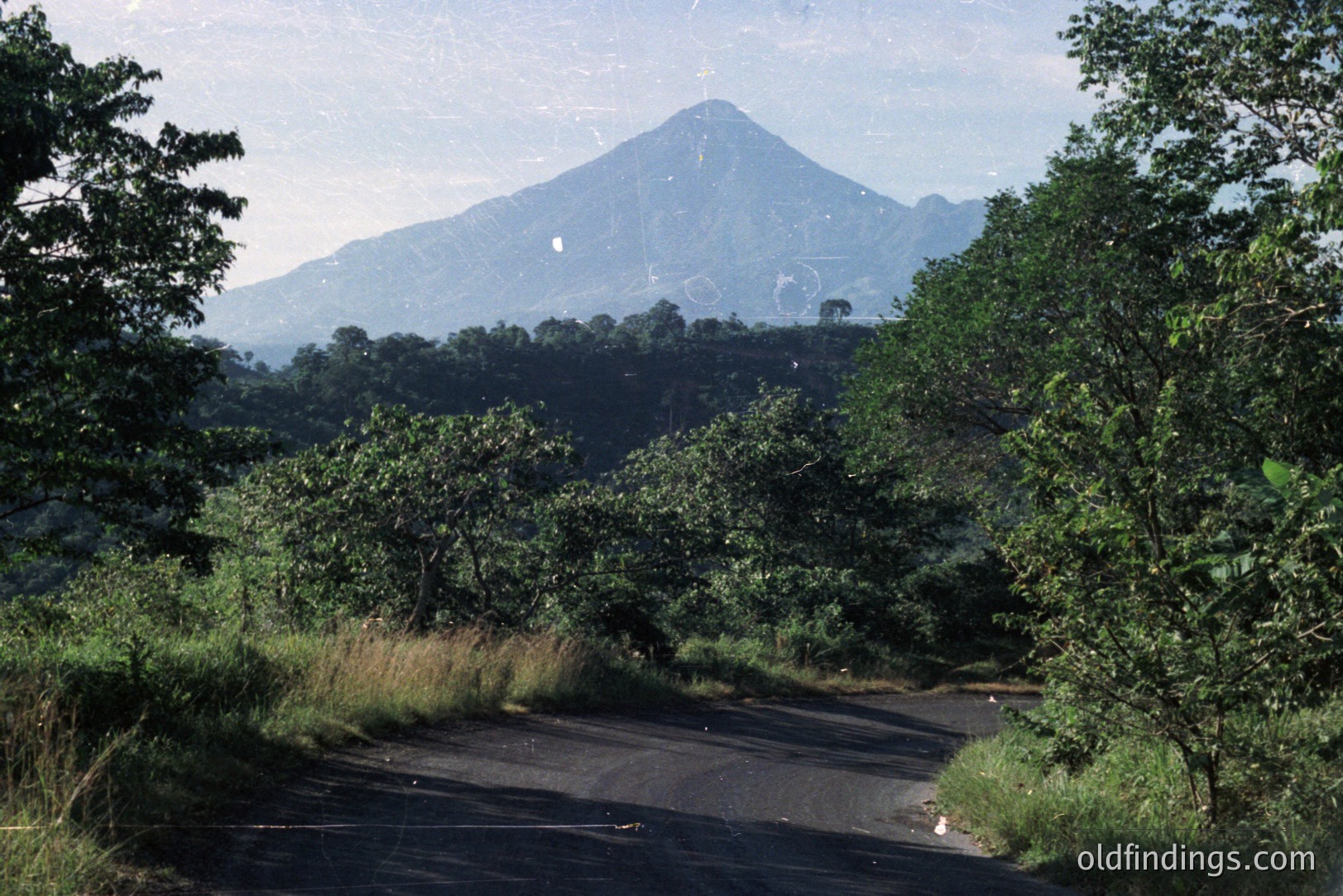 Lush tropical vegetation frames a winding asphalt road leading to a distinctive, cone-shaped volcano. Likely taken in Central America, possibly Costa Rica or Guatemala. The photograph’s faded colors suggest a 1970s or 1980s origin, possibly for travel or tourism documentation. Good for landscape design inspiration.