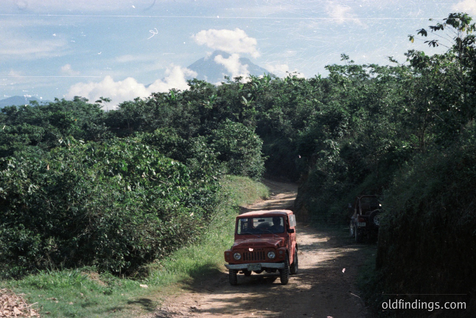 A red Fiat 500 Jeep navigates a narrow dirt road flanked by dense tropical foliage. A dormant volcano is visible in the distance, partially obscured by clouds. The photograph evokes a sense of exploration and adventure, likely from the 1970s or 80s. Potential stock image value for travel/adventure themes.