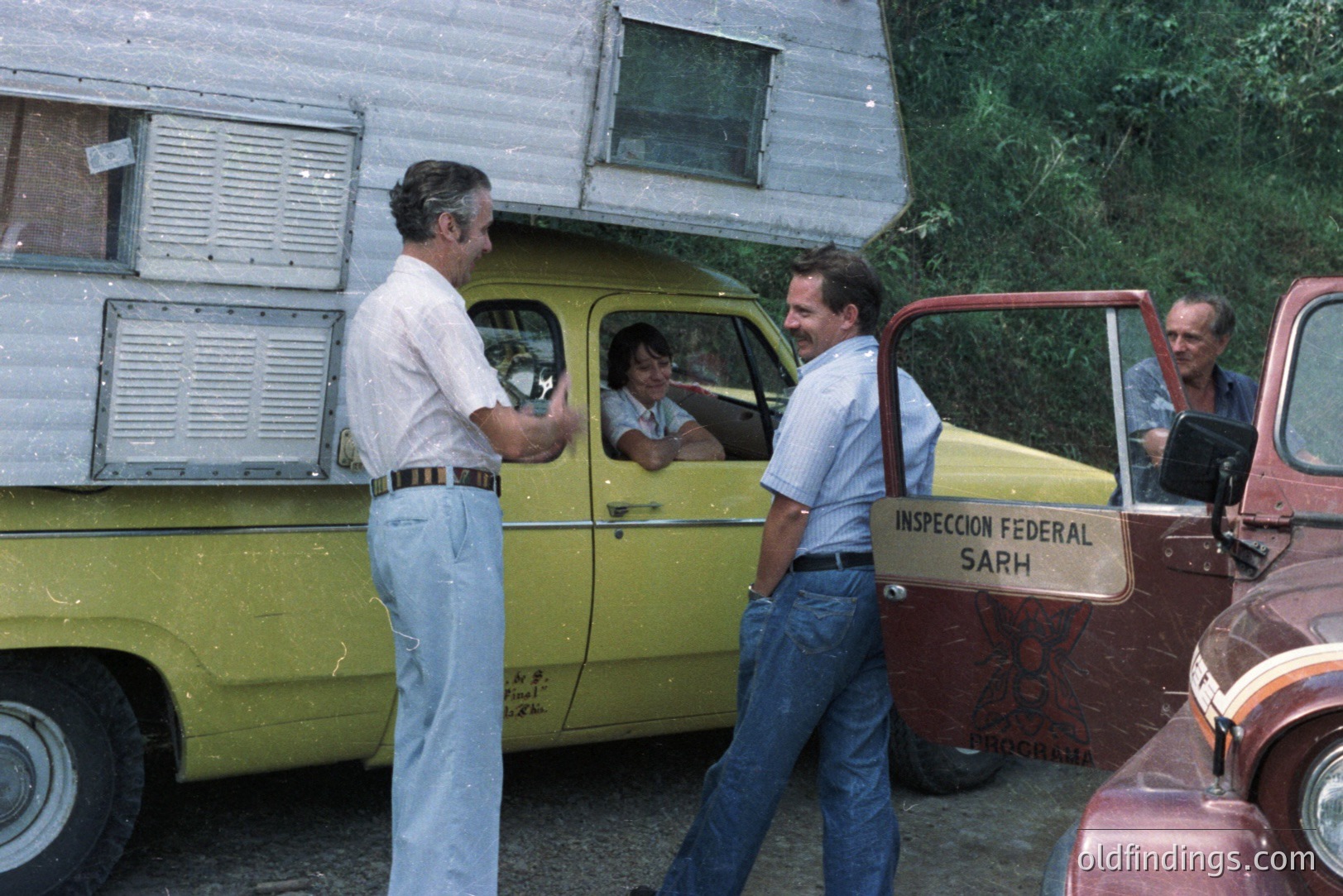 Two men in short sleeves converse beside a vintage yellow truck with a trailer. An older woman drives, and another man observes from the truck bed. "Inspección Federal SARH" placard visible. Likely 1960s-1970s, potentially Mexico based on signage. Classic truck details highlight automotive history.