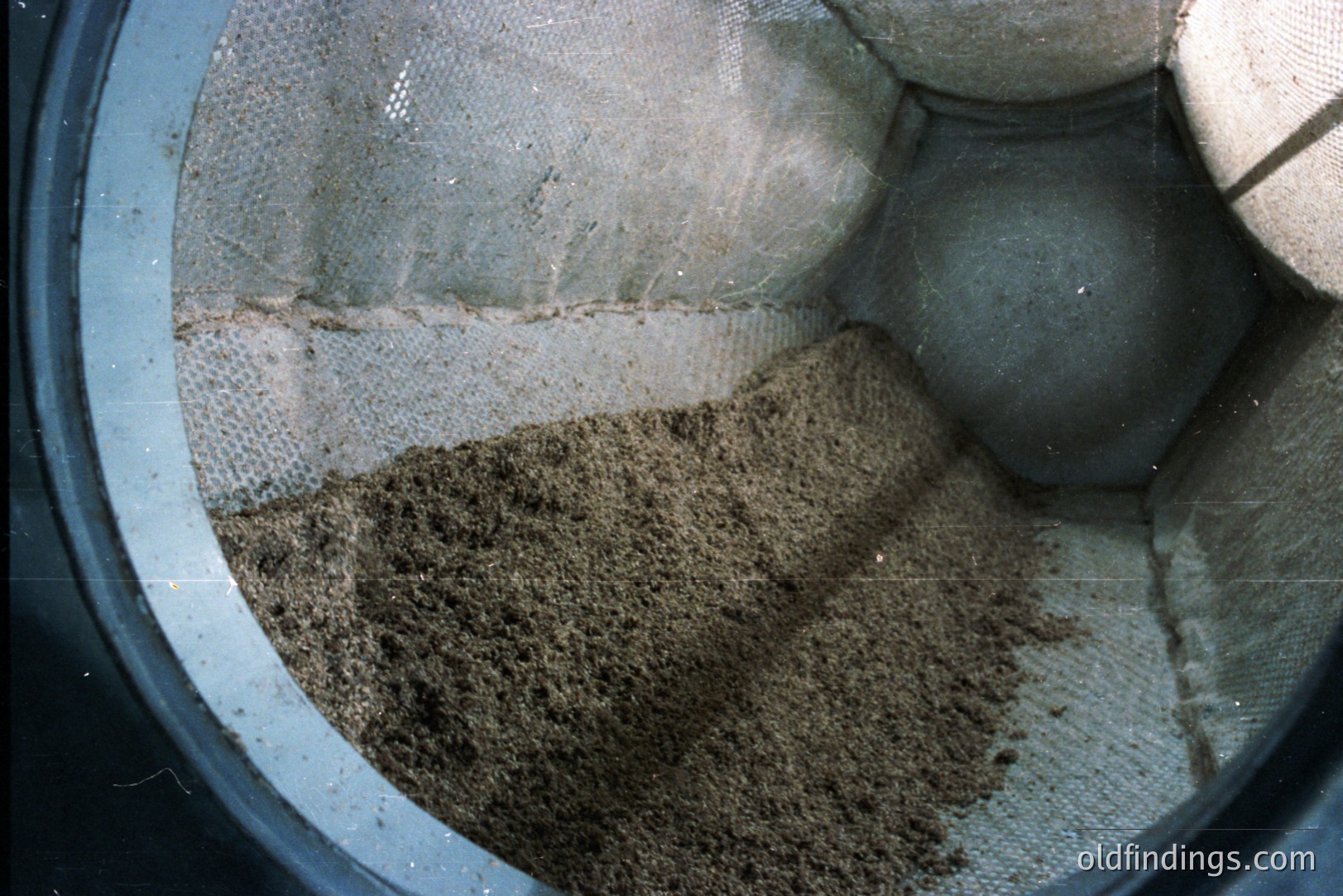 Close-up image of a textile mesh container filled with what appears to be dry, granular material, possibly soil or sand. The container's interior is lined with a light-colored fabric. The image has a slightly aged, muted color palette, suggesting a vintage or archival origin. Texture and material study potential.