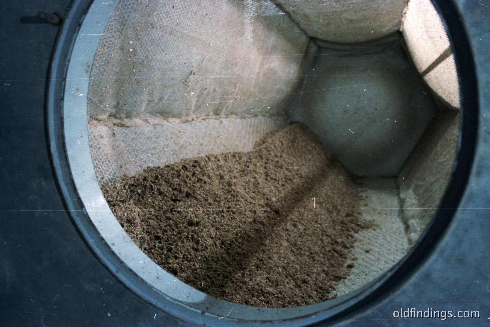 Close-up view within a dark, circular structure reveals layered sediment. Predominantly earthy browns and tans form a granular deposit, suggesting soil or mineral material. Textured, grainy surfaces fill the frame; faint light illuminates the interior. Appears to be a geological sample or construction site detail.