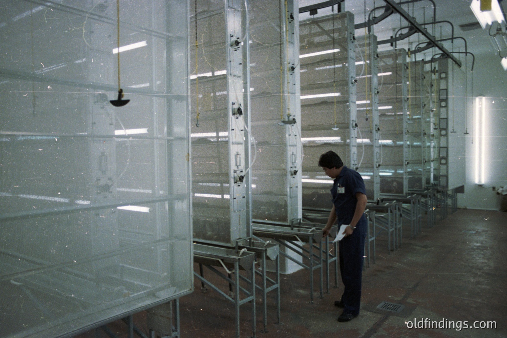 An industrial spray booth system, featuring multiple stainless steel panels suspended from overhead tracks. A worker in a dark blue uniform and dark pants examines a document, standing near a tray within the booth. Likely a manufacturing facility, possibly for metal products. Appears to be from the 1970s-1980s.