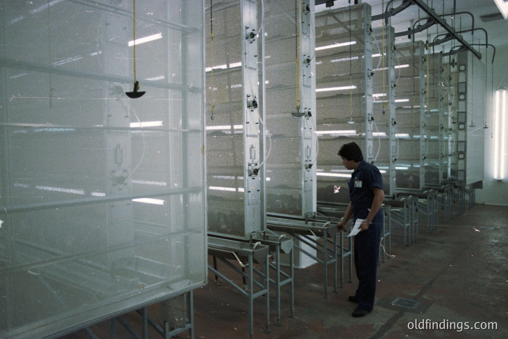 Industrial spray booth with a worker examining work in progress. Rows of metal frames with mesh screens suggest a painting or coating process. Fluorescent lighting and ventilation systems visible. Likely 1970s-80s manufacturing setting. Appears functional & utilitarian.