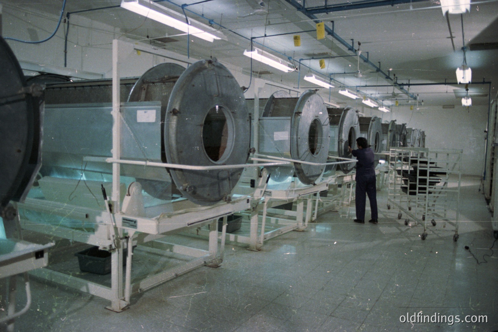 Industrial lab scene: multiple film processing tanks line a long room. A technician in dark clothing works alongside the machinery. Likely a film processing facility, potentially from the 1970s or 80s given the equipment's style. The lab's aesthetic could be useful for retro-themed design.