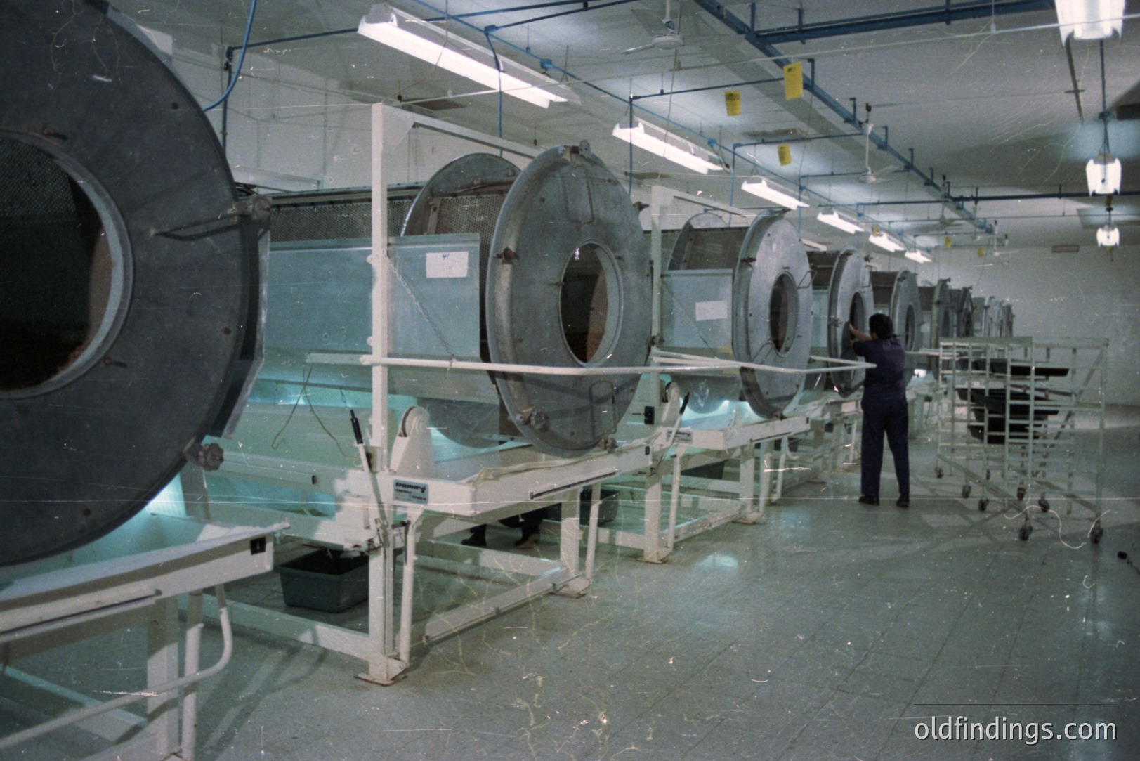Industrial view of a film processing facility. Multiple circular, stainless steel developing tanks line a brightly lit, cleanroom-like space. A lone technician is visible working on equipment. Likely 1970s or 1980s photographic lab. Appears unused/archival.