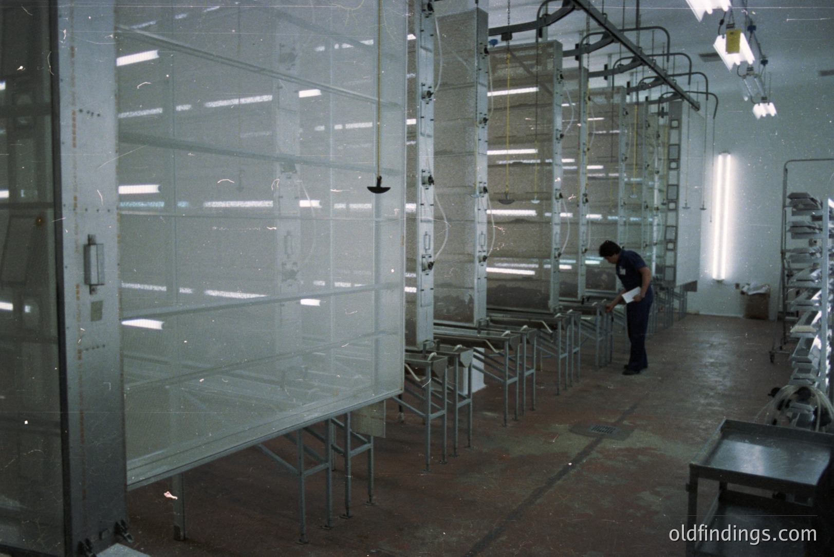Industrial scene: a worker inspects unfinished metal window frames on an automated conveyor system in a large spray booth. Overhead piping and lighting are visible. Likely a manufacturing facility, potentially 1970s-80s. Design reference for industrial processes.