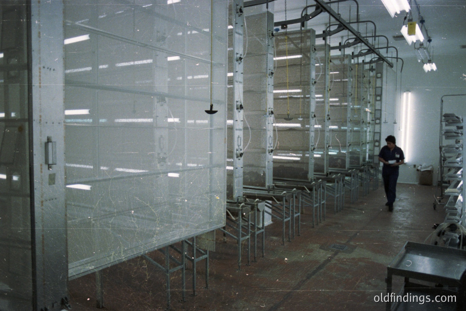 Industrial photograph showing a long, automated powder coating or spray painting line. Numerous racks with metallic components traverse the space under a network of overhead piping and lighting. A worker in overalls stands observing the process. Likely mid-to-late 20th century industrial setting.
