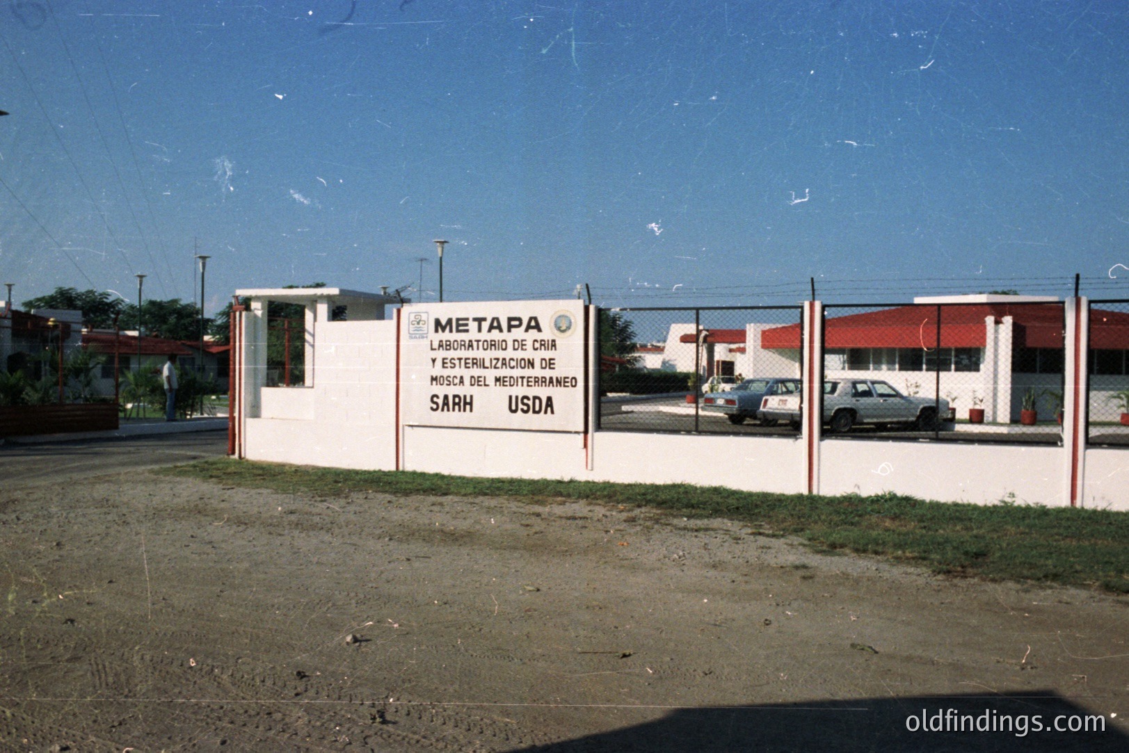Exterior view of the "Metapa" laboratory, breeding and sterilization of the Mediterranean fly. Sign indicates collaboration with the USDA. Institutional architecture, a red-roofed building is visible beyond a low fence. Possibly rural setting. 1980s aesthetic.