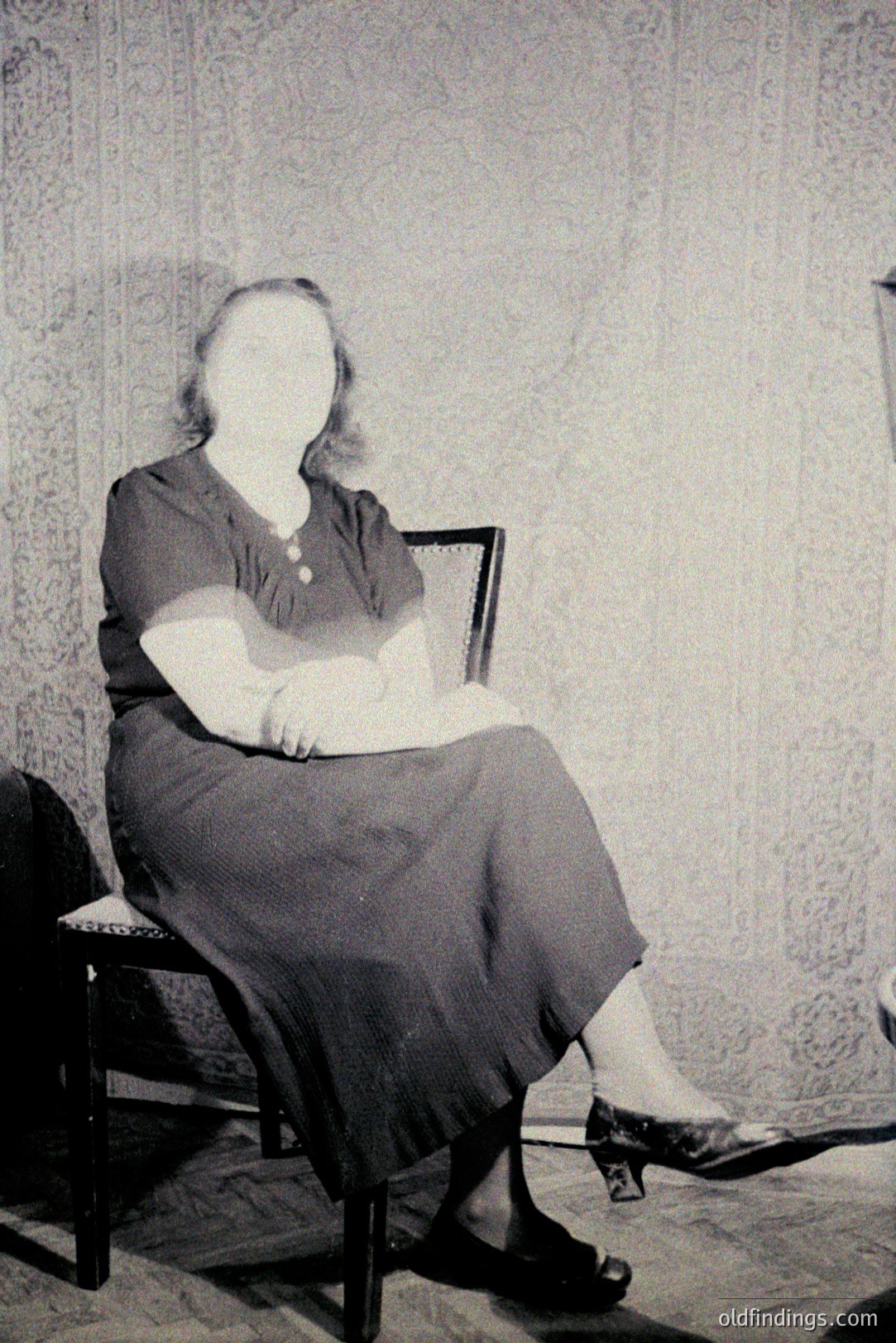 A formally posed, black and white portrait shows a woman seated in a dark chair. She wears a simple, long-sleeved dress with a buttoned bodice and a long skirt. The setting appears to be a home interior, with patterned wallpaper in the background. Likely 1930s-1950s. A study in domesticity and portraiture.