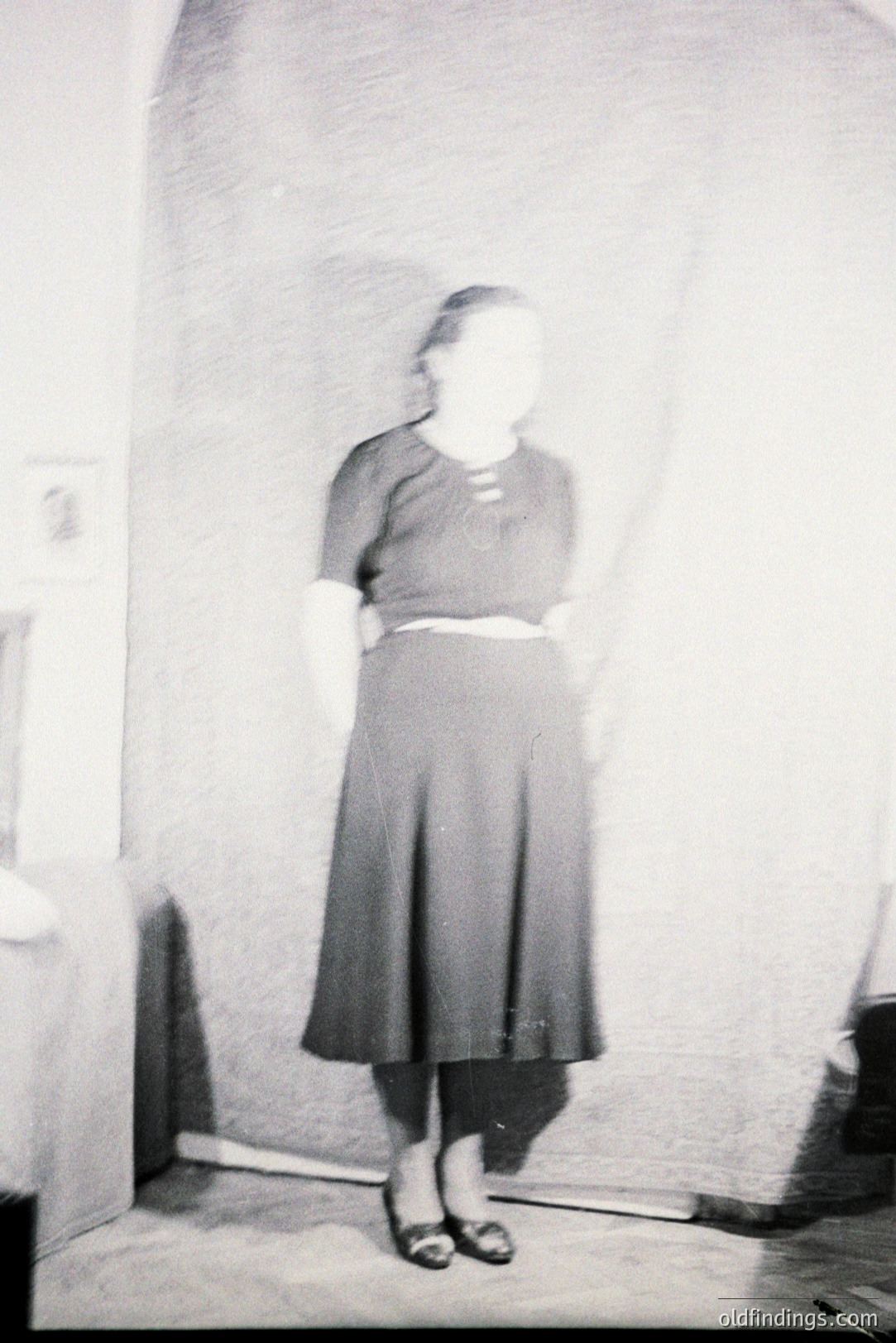 A full-length portrait depicts a woman in a mid-century style dress, featuring a fitted bodice and a full, pleated skirt. She wears dark flats and a white belt accentuates her waist. The background appears to be a textured brick wall. Likely 1950s fashion. Simple, classic style.