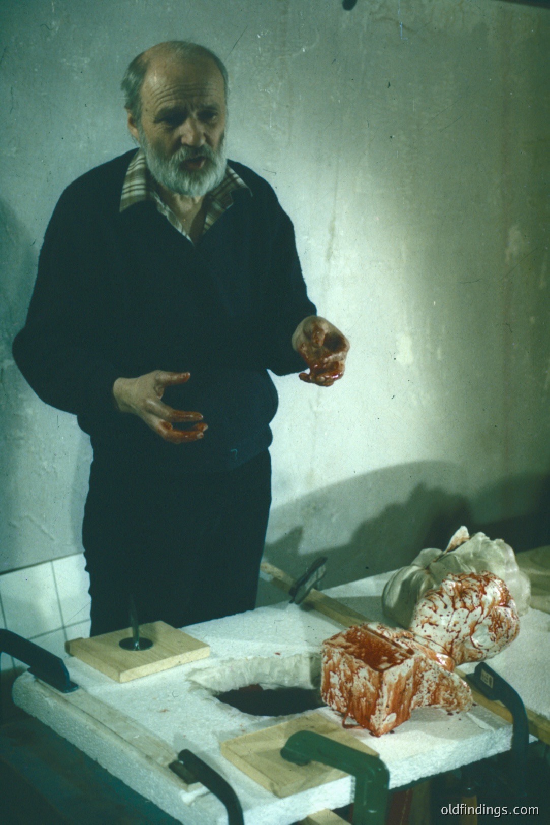 A middle-aged man with a full beard stands near a table holding a bloody, partially processed animal carcass. Likely a butcher or meat processor, he is wearing a dark sweater and appears to be demonstrating a cutting technique. The setting suggests a workshop or processing area.