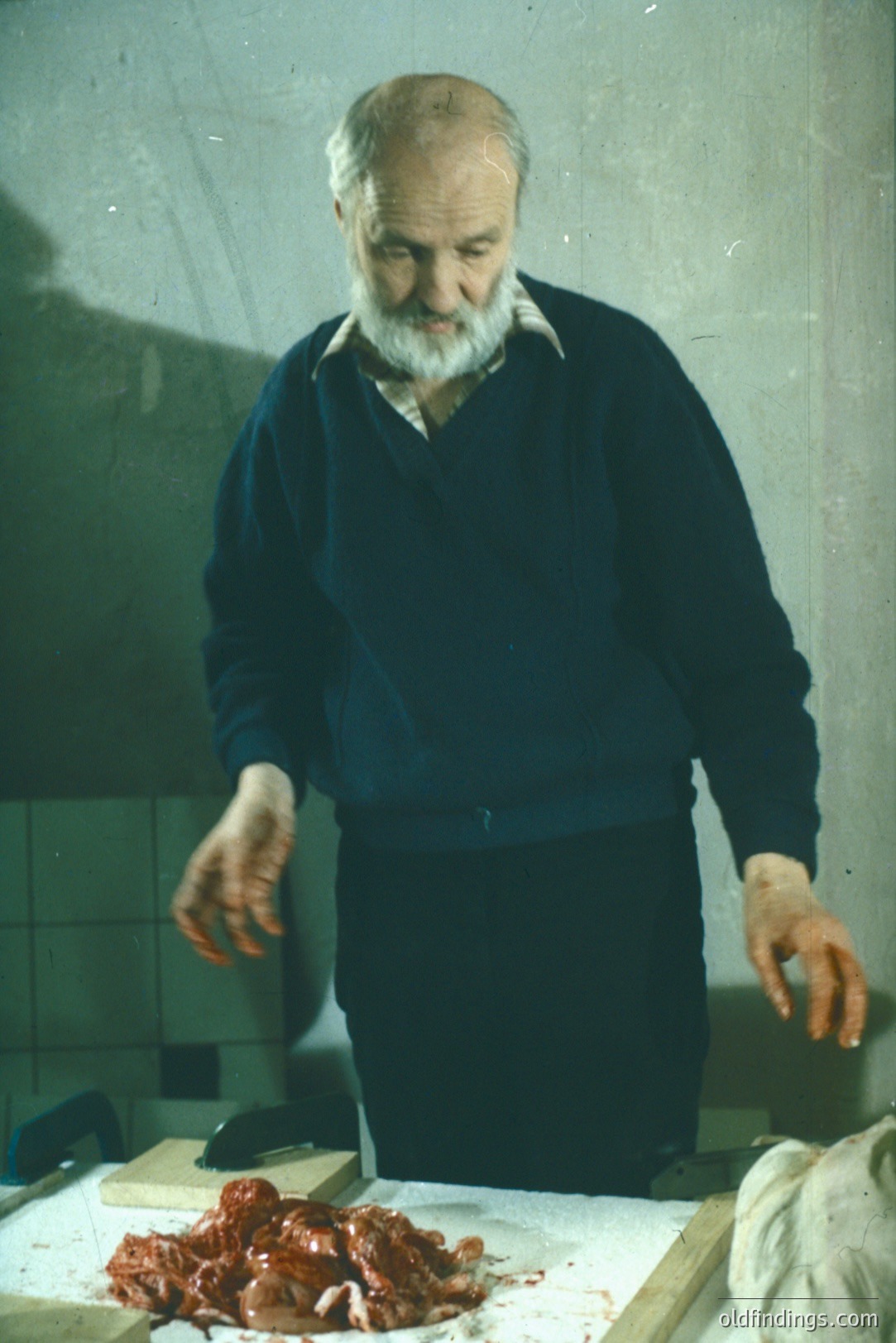 A butcher, likely middle-aged, stands behind a butcher block displaying cuts of raw meat. He wears a dark blue sweater and appears to be inspecting the meat. The setting suggests a small, older shop, with tiled walls and basic equipment. Likely 1960s-1970s.