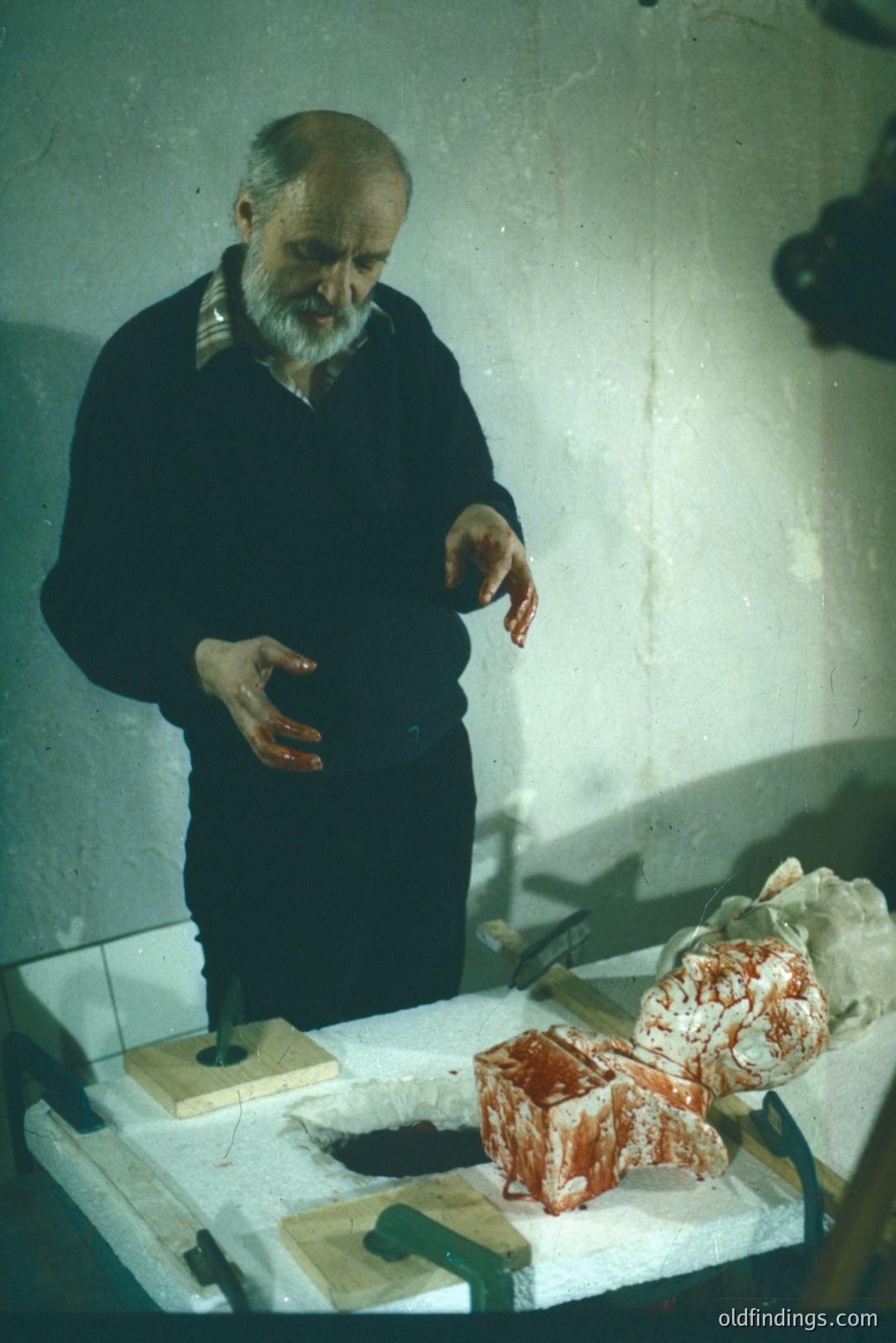 A butcher, with a gray beard and dark sweater, stands at a stainless-steel table. He appears to be examining a large piece of meat. Visible are various tools and cuts of red meat, including a rectangular block and a marbled cut. Likely a vintage photo from the mid-20th century, potentially demonstrating a traditional meat preparation process.