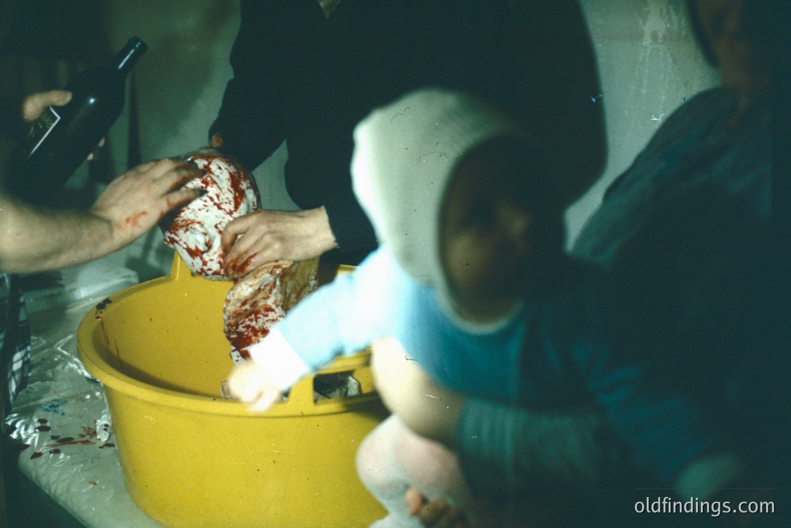 A close-up shows hands in latex gloves working with a large portion of raw, intensely red meat in a yellow plastic tub. A child in a blue outfit observes from the foreground. The scene appears domestic, possibly a food preparation or butchery process. Appears to be a 1970s or 80s snapshot.