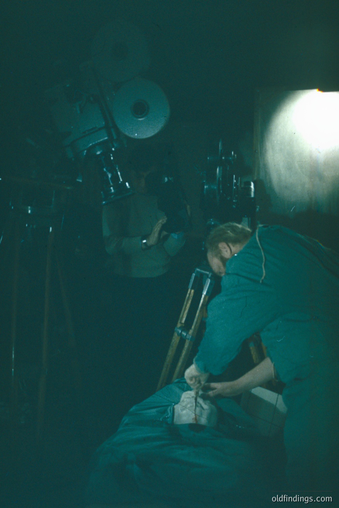 A figure in a teal work shirt is crouched, adjusting a vintage camera mounted on a tripod. Large, older studio lighting equipment is visible in the background, lit with a bright, focused lamp. Likely a behind-the-scenes studio shot, possibly from the 1970s or 80s, capturing an analog photography workflow.