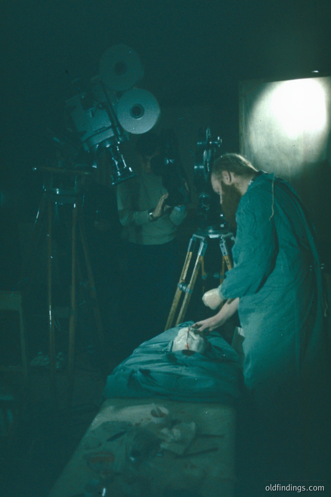 A man in a green surgical gown leans over a figure covered by a sheet on a bed. Elaborate stage lighting equipment, including large reflectors, is visible in the background. Likely a film or theater production still. The image conveys a clinical, unsettling atmosphere.