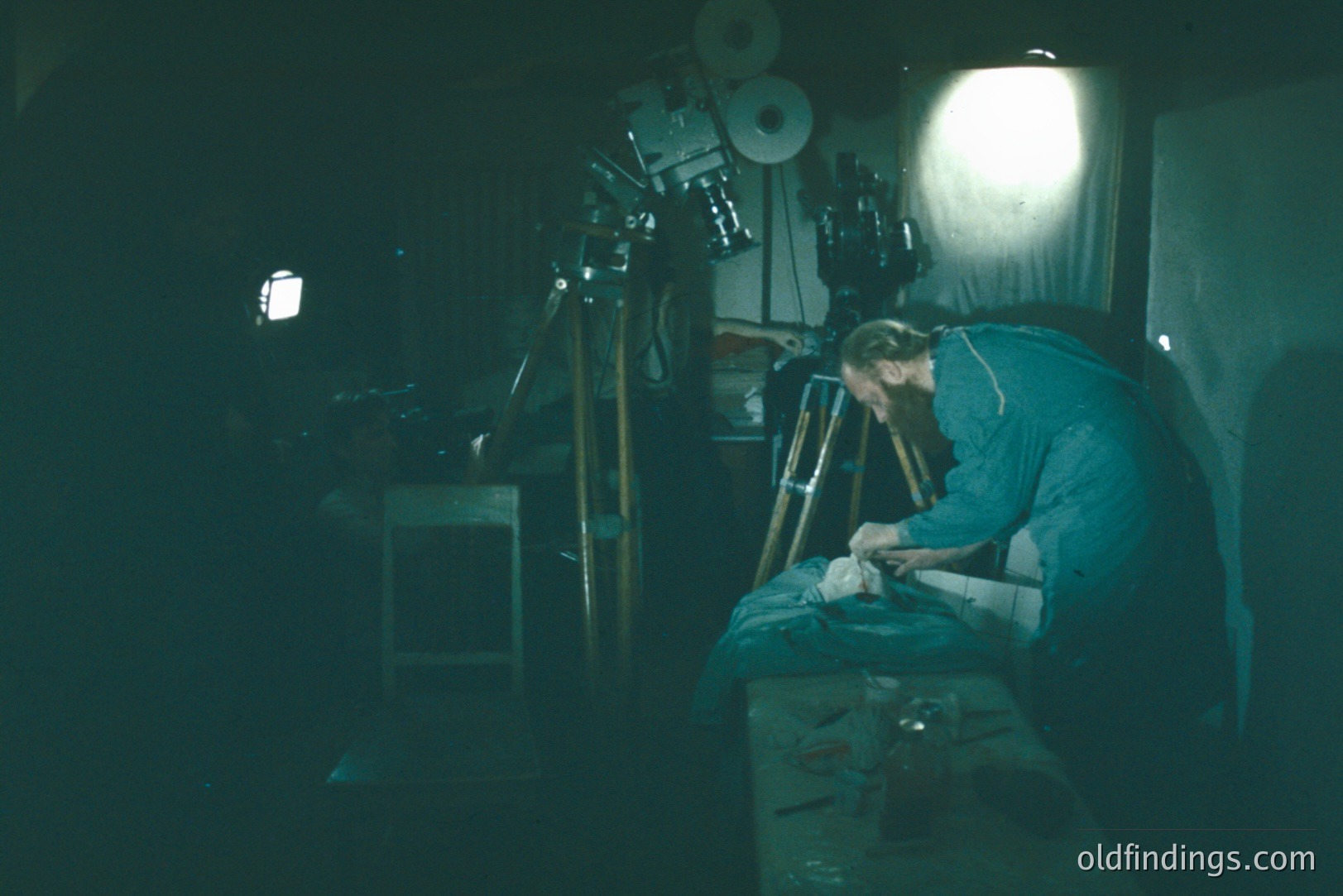 A man in a blue work shirt meticulously repairs a film reel in a dimly lit, cluttered workspace. Visible are film spools, tools, and a tripod-mounted camera, suggesting a film processing or repair environment. Likely mid-20th century, possibly 1950s-1970s.