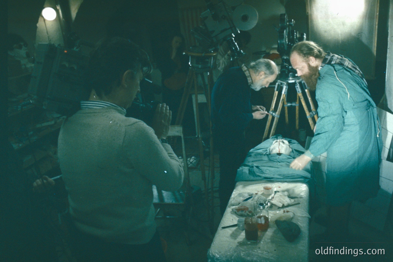 Three figures are depicted in a dimly lit, cluttered workshop. A camera operator adjusts equipment, while two others meticulously examine what appears to be a taxidermied animal on a table. Tripods and lighting gear surround them. Appears to be a 1970s studio environment.