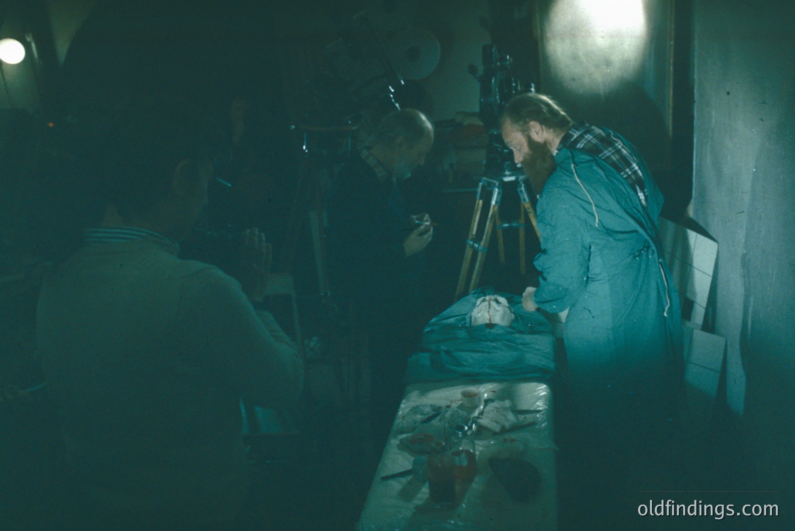 A photograph depicting three figures in a dimly lit space, likely a darkroom. Two men examine a developed photograph laid on a table; one adjusts a tripod. The setting appears utilitarian, with visible equipment and a sense of focused work. Evokes vintage photography processes.