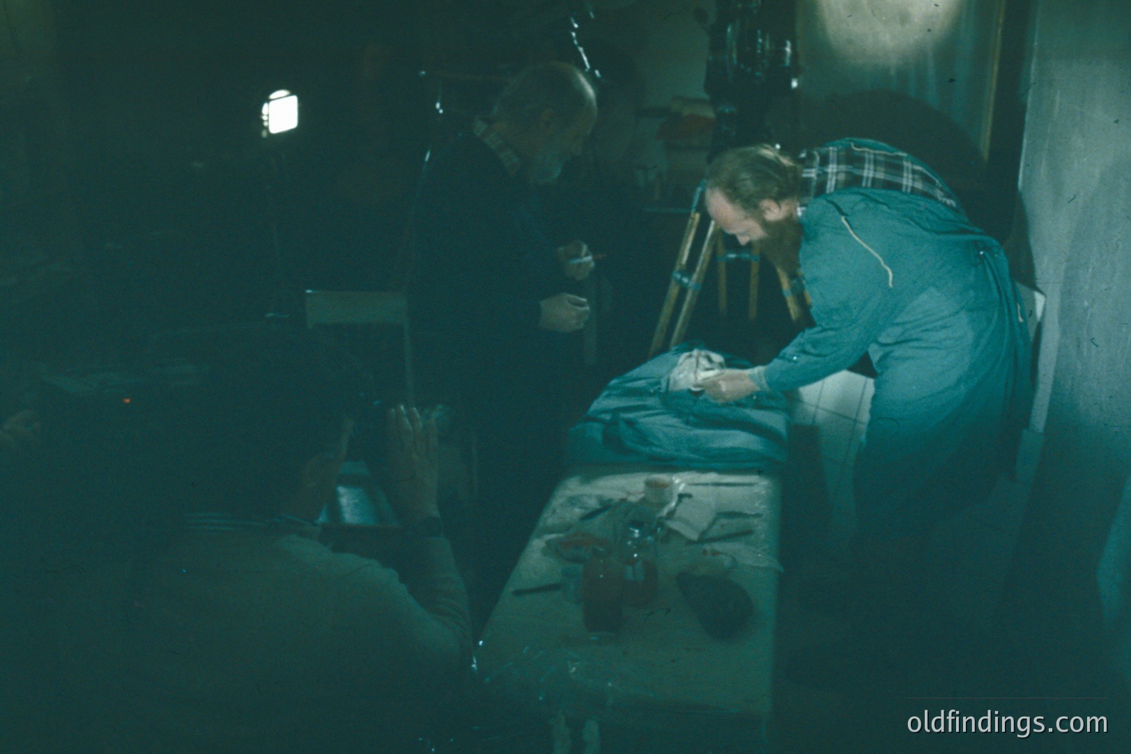 A man with a beard examines what appears to be a bird specimen on a table, while another figure observes. The scene is dimly lit, suggesting a lab or workshop setting. Visible equipment includes bottles, tools, and a tripod. Likely mid-20th century, potentially scientific documentation.