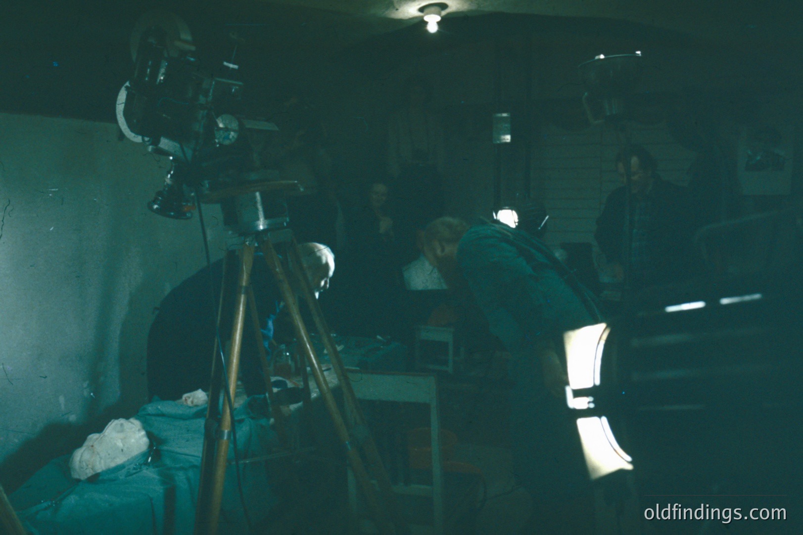 A patient lies under a camera rig, likely for a medical or scientific procedure, in what appears to be a basic, institutional setting. Three figures are visible, seemingly observing or assisting. The tripod and lighting indicate a deliberate, controlled environment. Mid-20th century aesthetic.