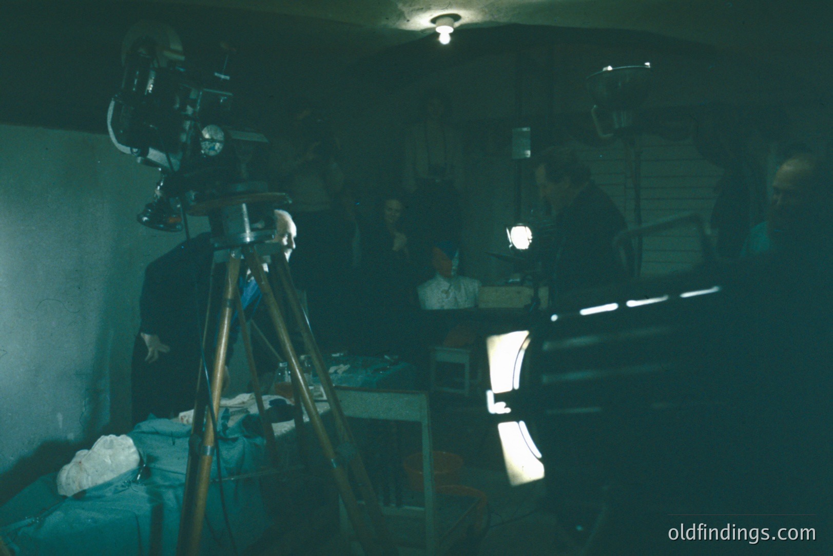 A film set scene: a vintage 16mm camera mounted on a tripod dominates the foreground. Figures are partially visible in shadow, engaged in what appears to be filming activity, likely a documentary or experimental project. Low lighting and stark contrasts create a film noir aesthetic.