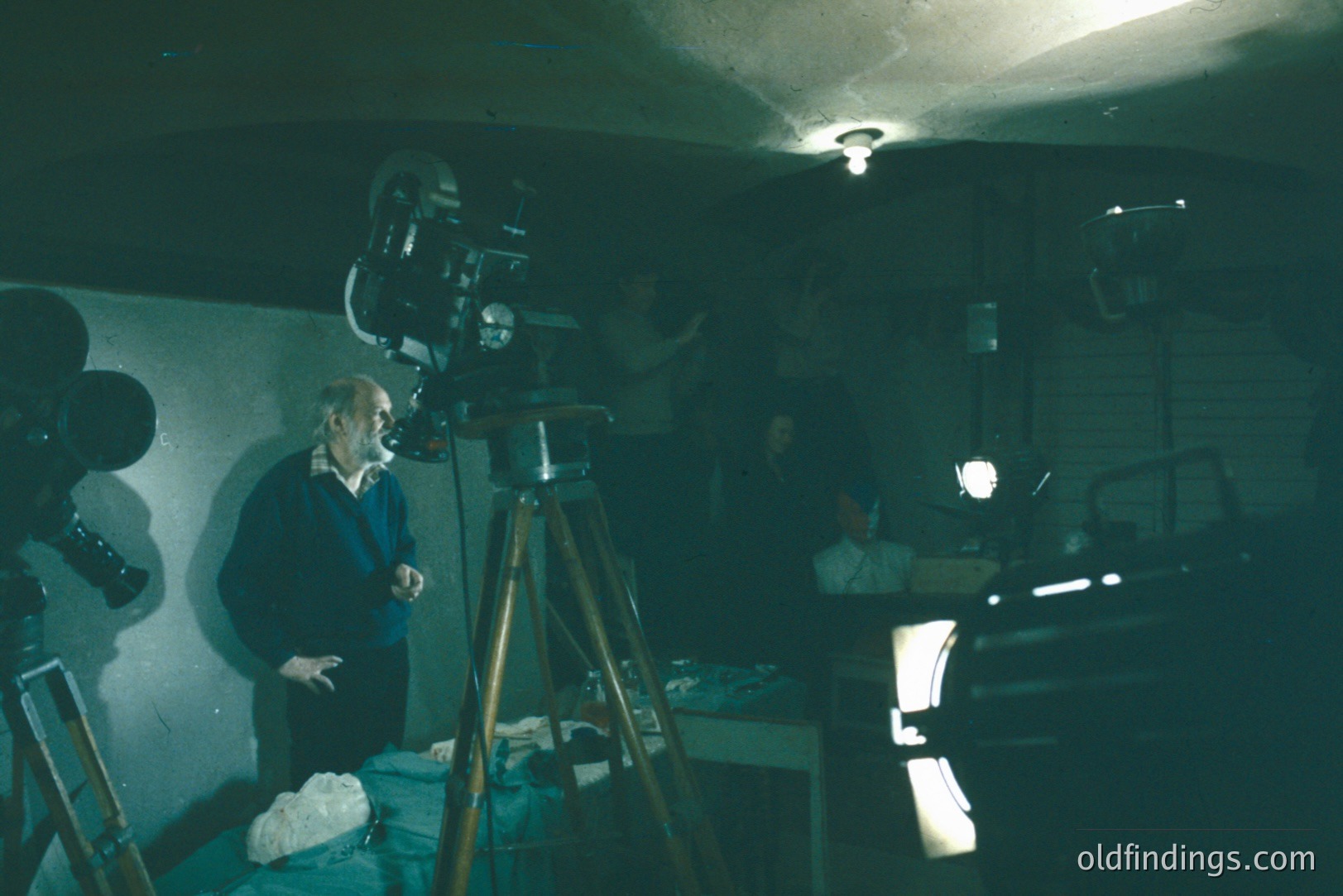 A film set interior, likely a studio or basement, features a cameraman adjusting a vintage film camera on a tripod. Visible crew members in the background operate lights. Appears to be a 1960s-70s production. Potential for historical film industry research.