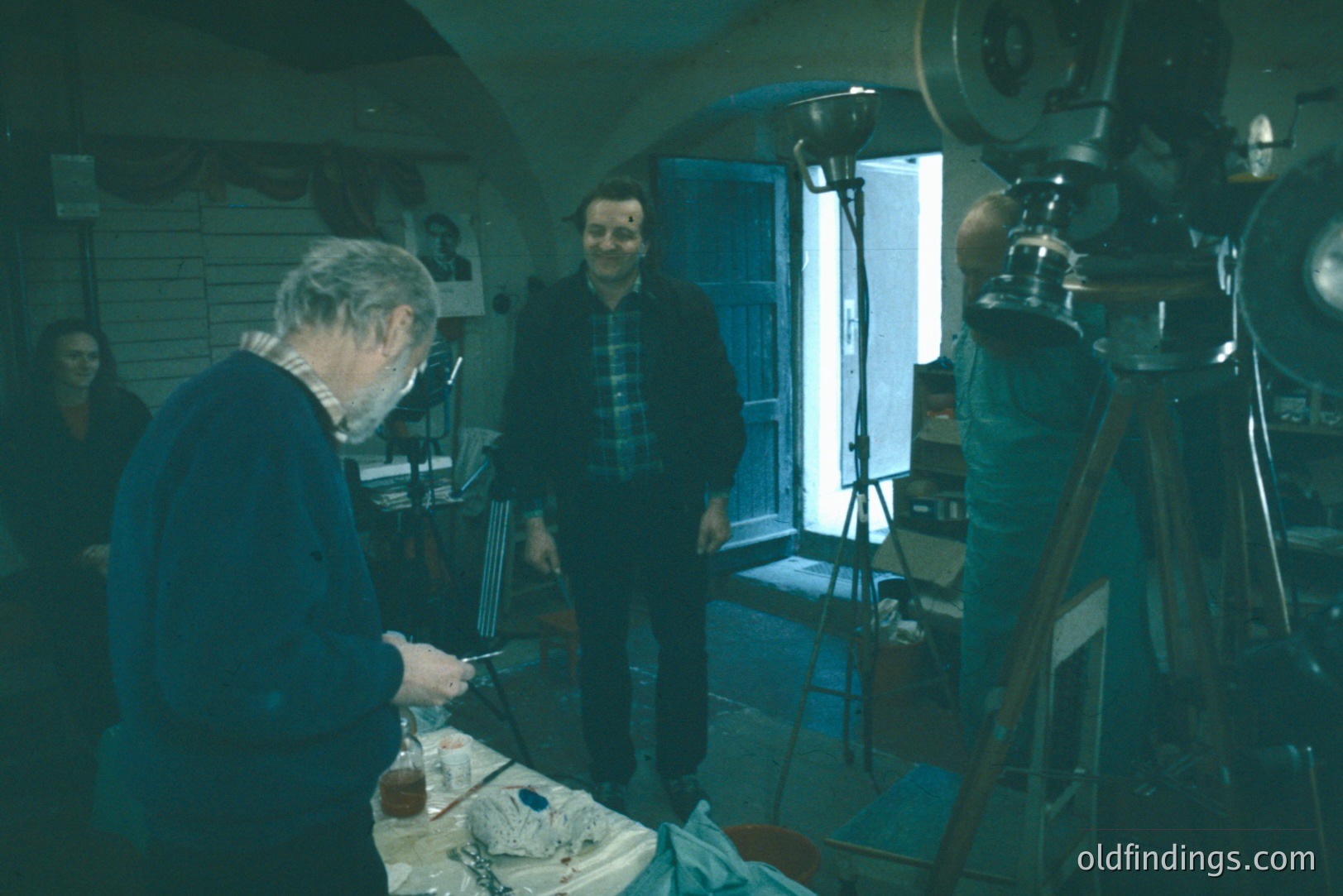 A man with a focused expression meticulously works on a sculpture in a cluttered studio. A second man in a plaid shirt stands near a window, observing. A large camera on a tripod sits in the corner. Likely a 1970s artistic workspace.