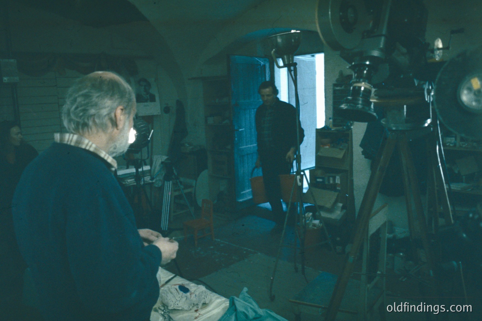 An older man, seated and working on a sculpture, is the focus in a cluttered, dimly-lit workshop. A large-format camera and lighting equipment occupy the foreground. A second person stands near a doorway, visible in the background. Likely a studio or artist's workspace, possibly 1970s.