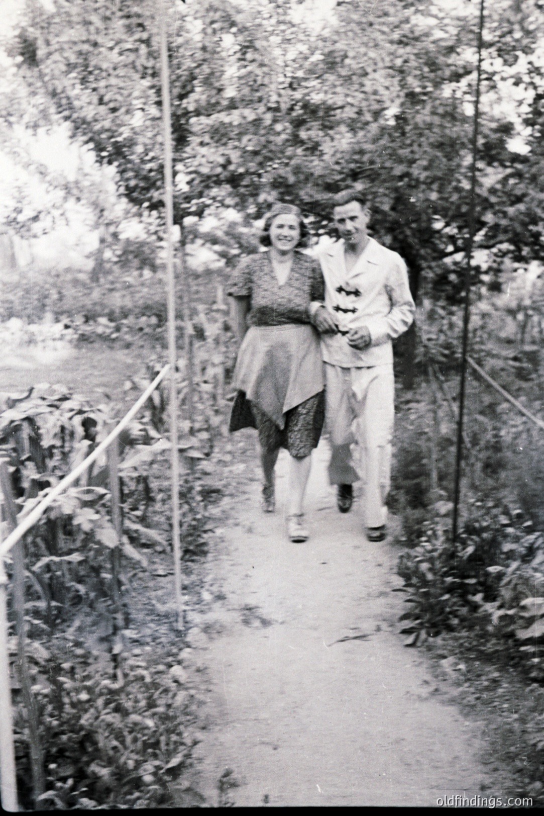 A couple strolls along a narrow, gravel path lined with vegetation. The woman wears a V-neck dress with a prominent sash; the man is in a light-colored suit with a bow tie. Likely a garden or park setting, c. 1930s-1950s. A glimpse into mid-century leisure.