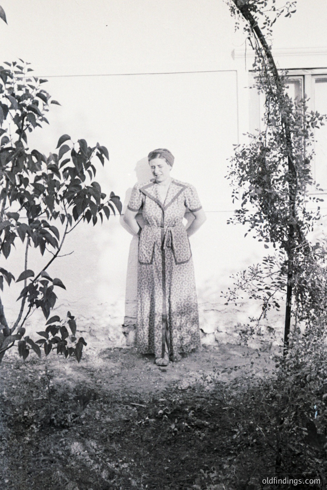 Formal portrait of a woman in a patterned, high-waisted dress, posed outdoors against a white wall & overgrown trellis. Appears to be a garden or courtyard setting. Likely 1930s-1950s era fashion. Could be valuable for historical costume reference.