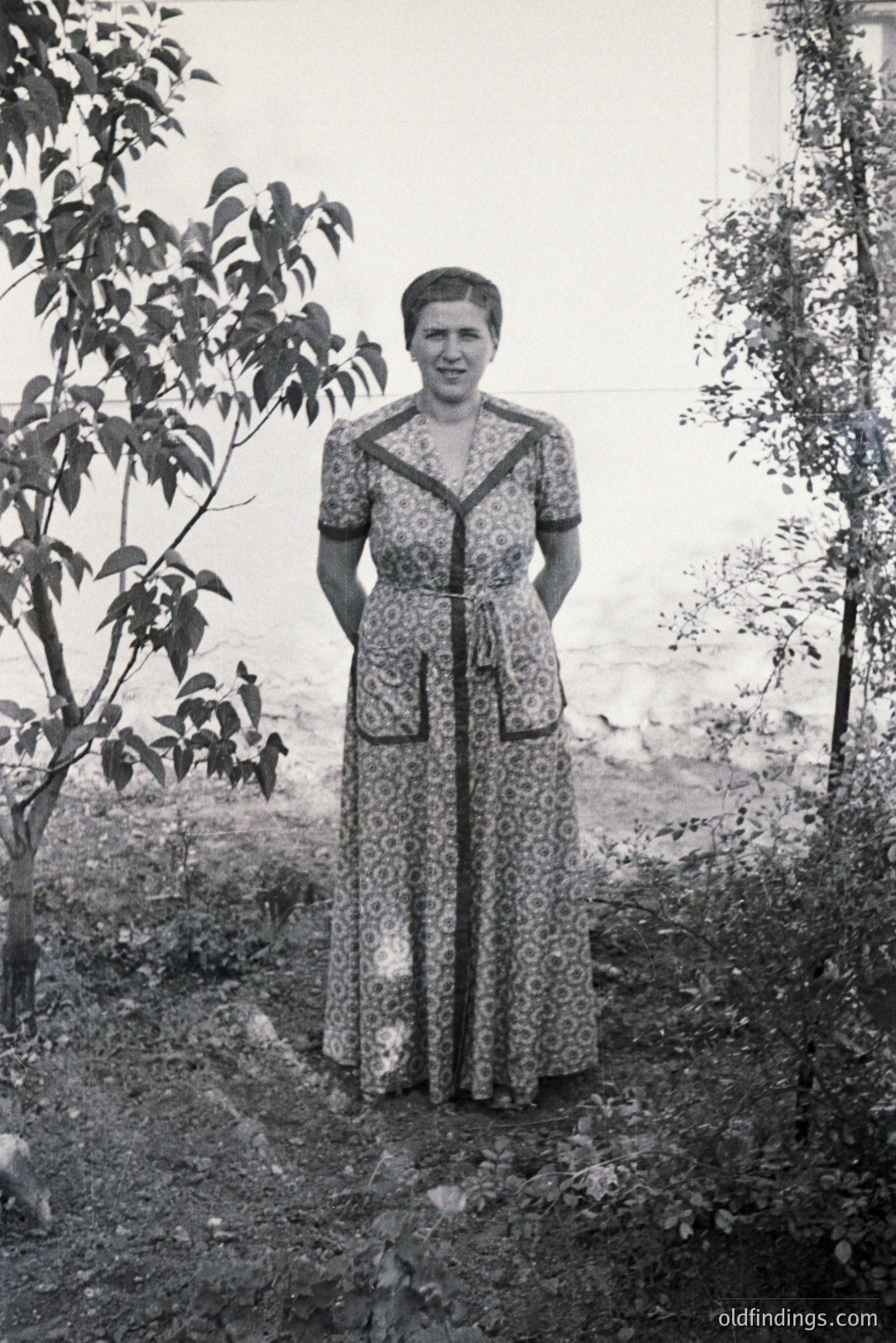 Full-length portrait of a woman in a long-sleeved, patterned dress, standing outdoors. Lush greenery and a stone wall form the backdrop. Likely taken in the mid-20th century, showcasing classic 1960s style. The dress's design and pose reflect the era's aesthetic.