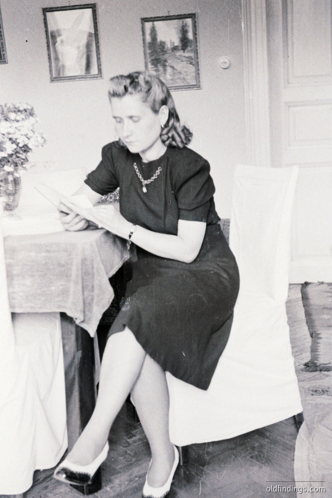 Woman seated at a table, examining papers, within an interior space framed by landscape artwork. She wears a dark dress, pearl necklace, and stylish heels. Likely a domestic or studio portrait from the 1940s. Offers insight into fashion & interior design of the era.