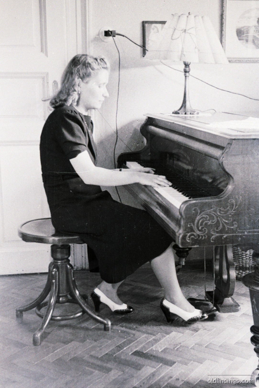A woman in a dark dress and pumps sits at an ornate, upright piano. Interior view shows a well-appointed room with hardwood floors & a table lamp. The scene suggests a domestic setting, likely 1940s-1960s. Architectural details denote European influence.