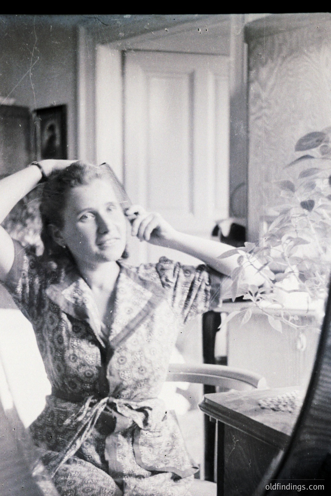 A seated woman in a patterned dress poses with her hands raised, appearing to adjust her hair. Interior scene with a patterned wallpaper and dark wood furniture. Likely a domestic setting, possibly a studio portrait. Appears to be a mid-20th century photograph, ~1940s-1950s.