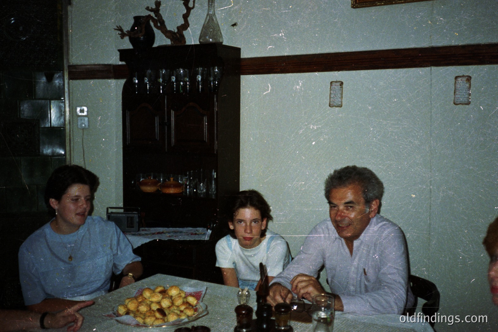 Three people sit at a table laden with pastries, likely a celebratory meal. The interior features a dark wood sideboard and paneled walls, suggestive of a mid-century aesthetic. The subjects appear to be a multigenerational family. Photo style implies a casual, domestic snapshot.