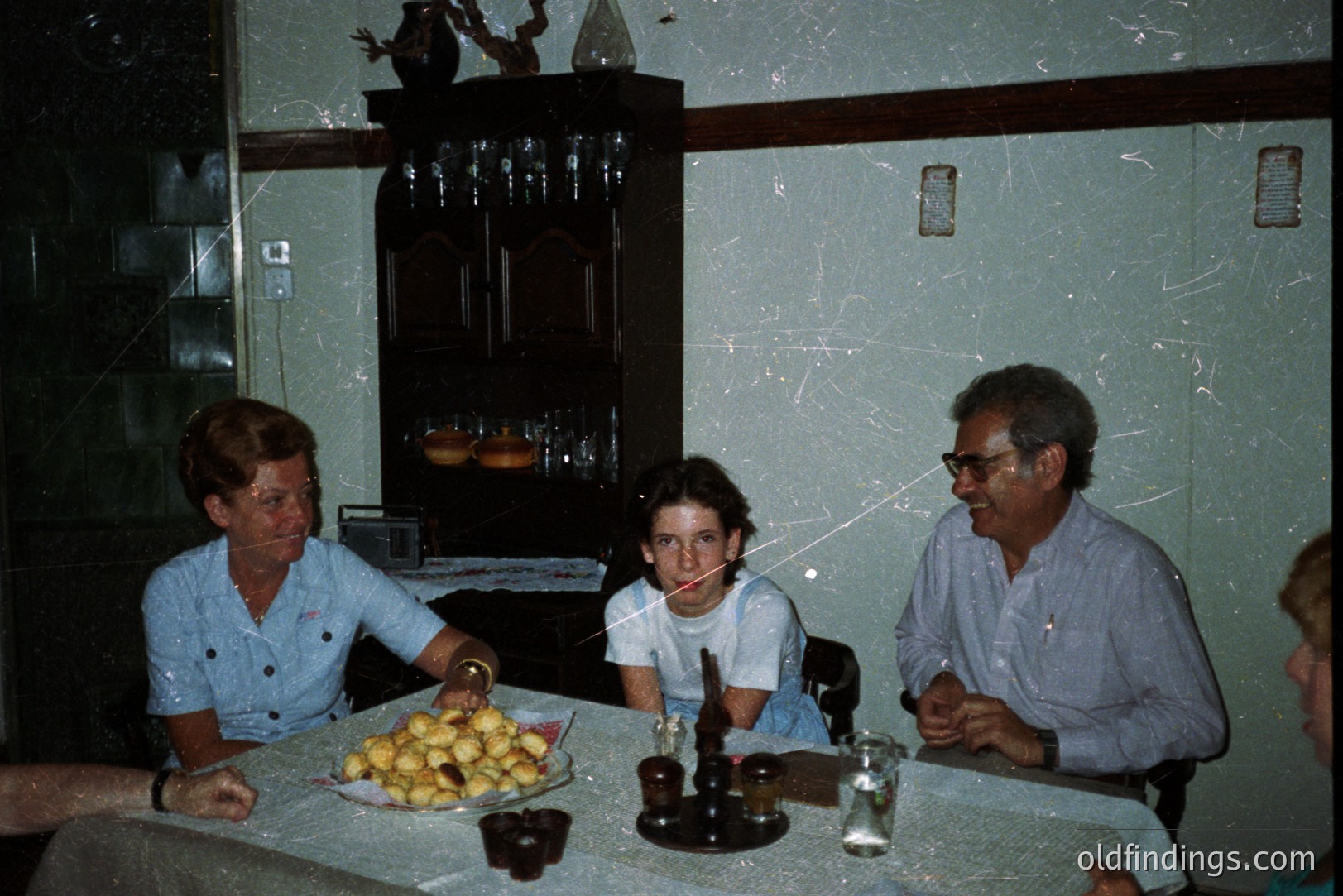 A family gathers around a table laden with food, likely enjoying a meal. Three individuals are visible – a woman in a blue shirt, a teenage boy, and an older man with glasses. The room's decor, featuring a dark wood cabinet and patterned wallpaper, suggests a 1970s domestic setting. A chess set rests on the tablecloth.