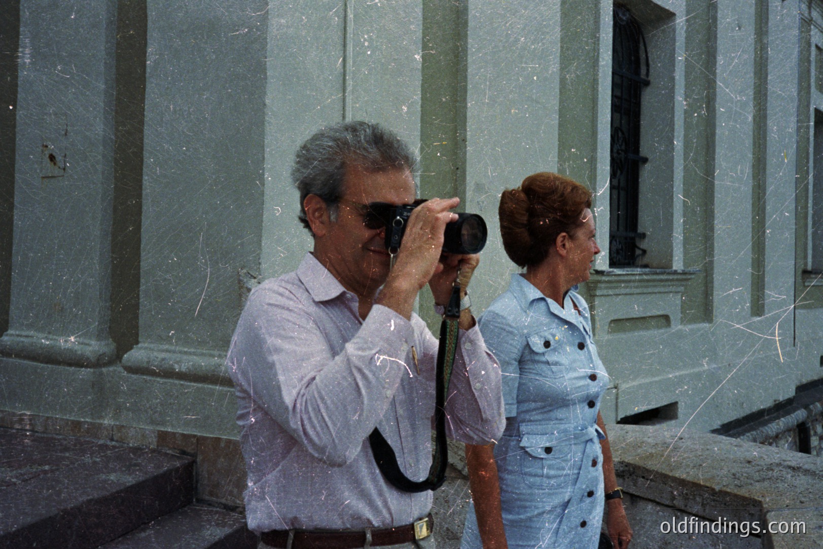 Man with a camera captures a moment alongside a woman in a 1970s-style blue dress. Architectural facade with classical details provides a backdrop. Likely a tourist snapshot showcasing travel and lifestyle. Visible film grain characteristic of the era. Location and photographer unknown.