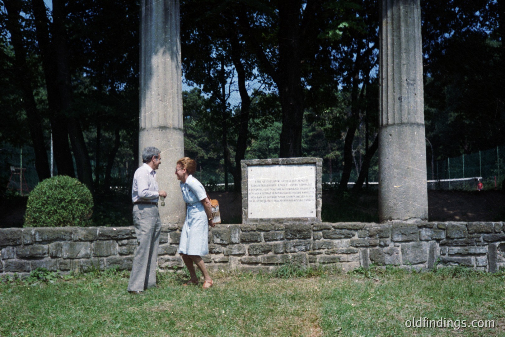 A man and woman, likely tourists, engage in conversation near a monument featuring two classical columns and a stone plaque. The architectural elements suggest a memorial or historical site. Location appears to be a park or formal garden. Clothing styles indicate a 1970s timeframe. A stone wall and greenery define the scene.
