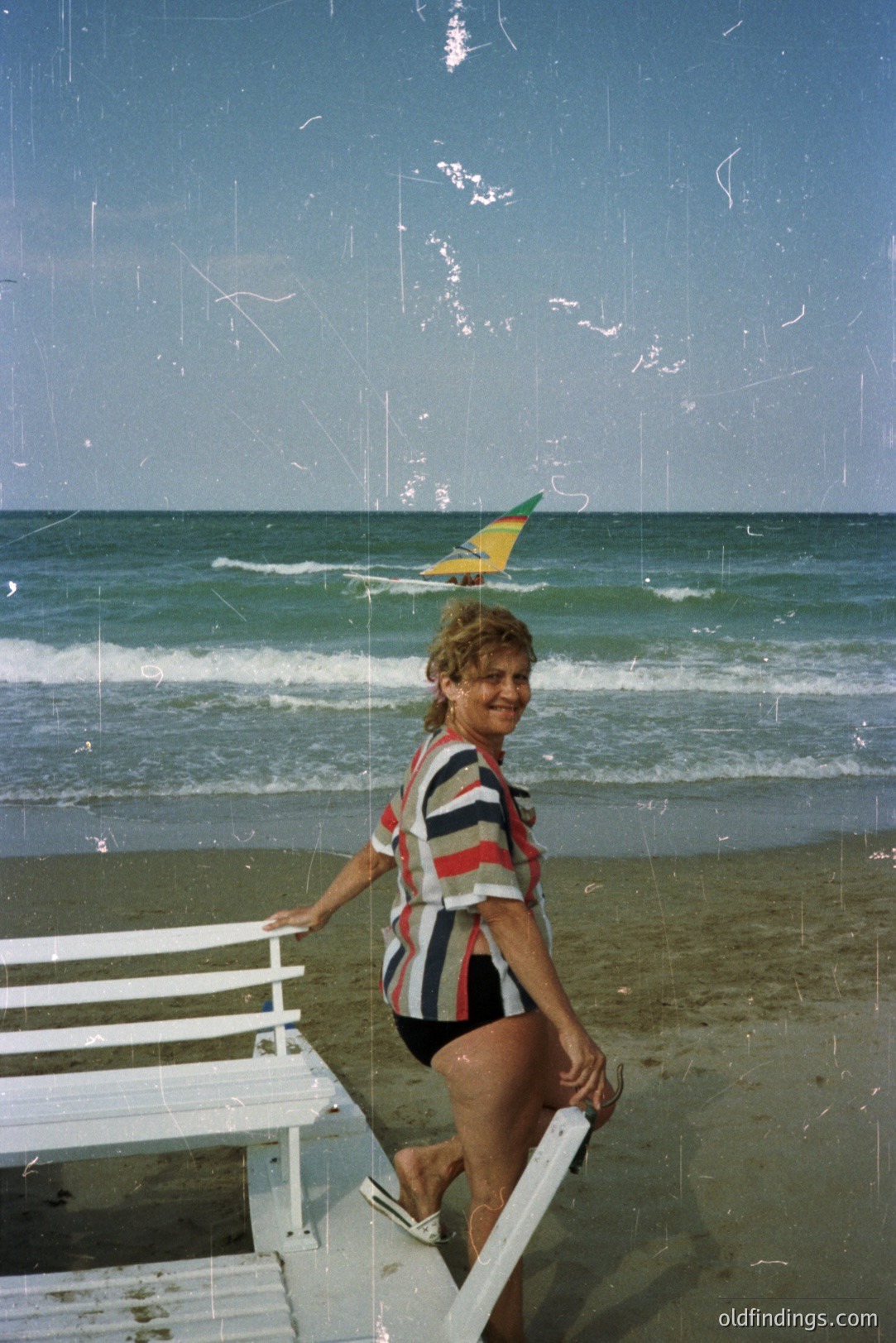 A woman with curly hair smiles on a beach, poised on a white metal lifeguard stand. She wears a striped athletic top & black shorts. A kitesurfer is visible in the background alongside the turquoise sea. Likely 1970s-80s, potentially Bulgaria's Black Sea coast. Vintage film grain is apparent.